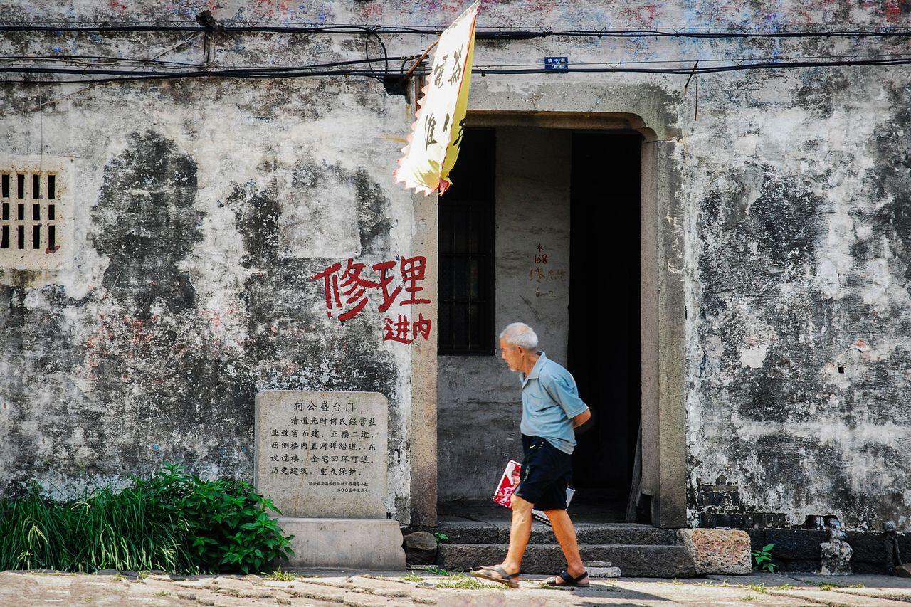 homme age chinois passant devant un batiment delabre
