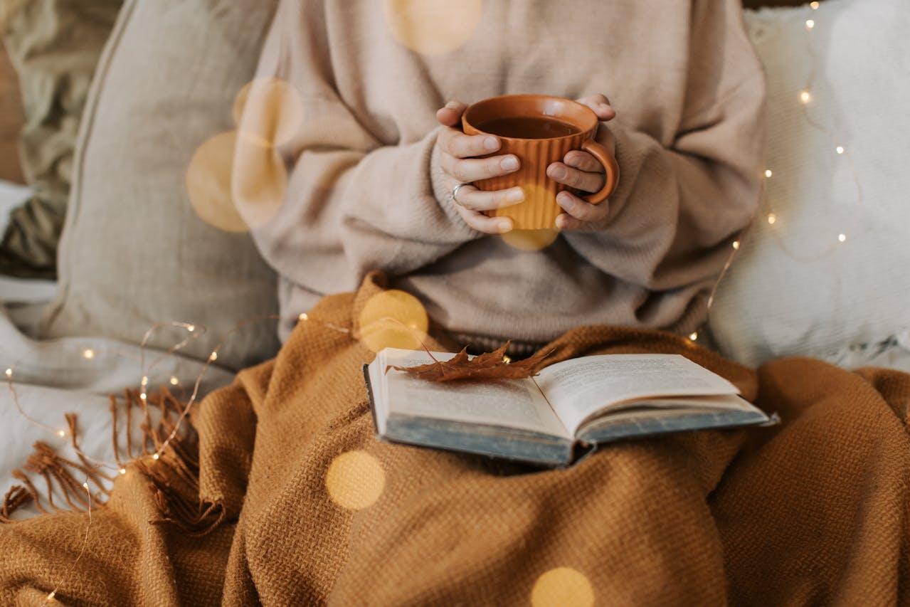 personne avec un plaid et du the lisant un livre sur un fauteuil