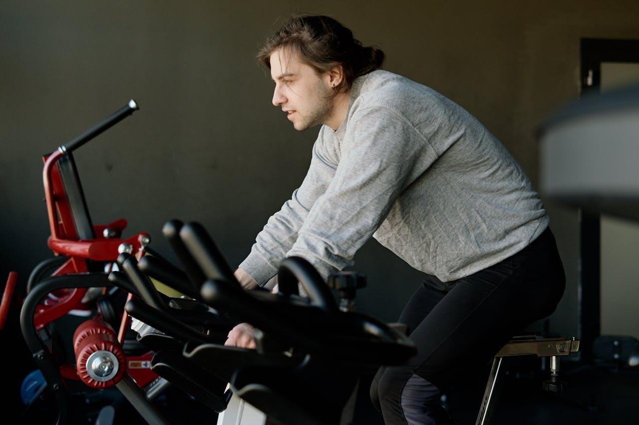 jeune homme en sweat gris pratiquant le velo spinning en salle de sport
