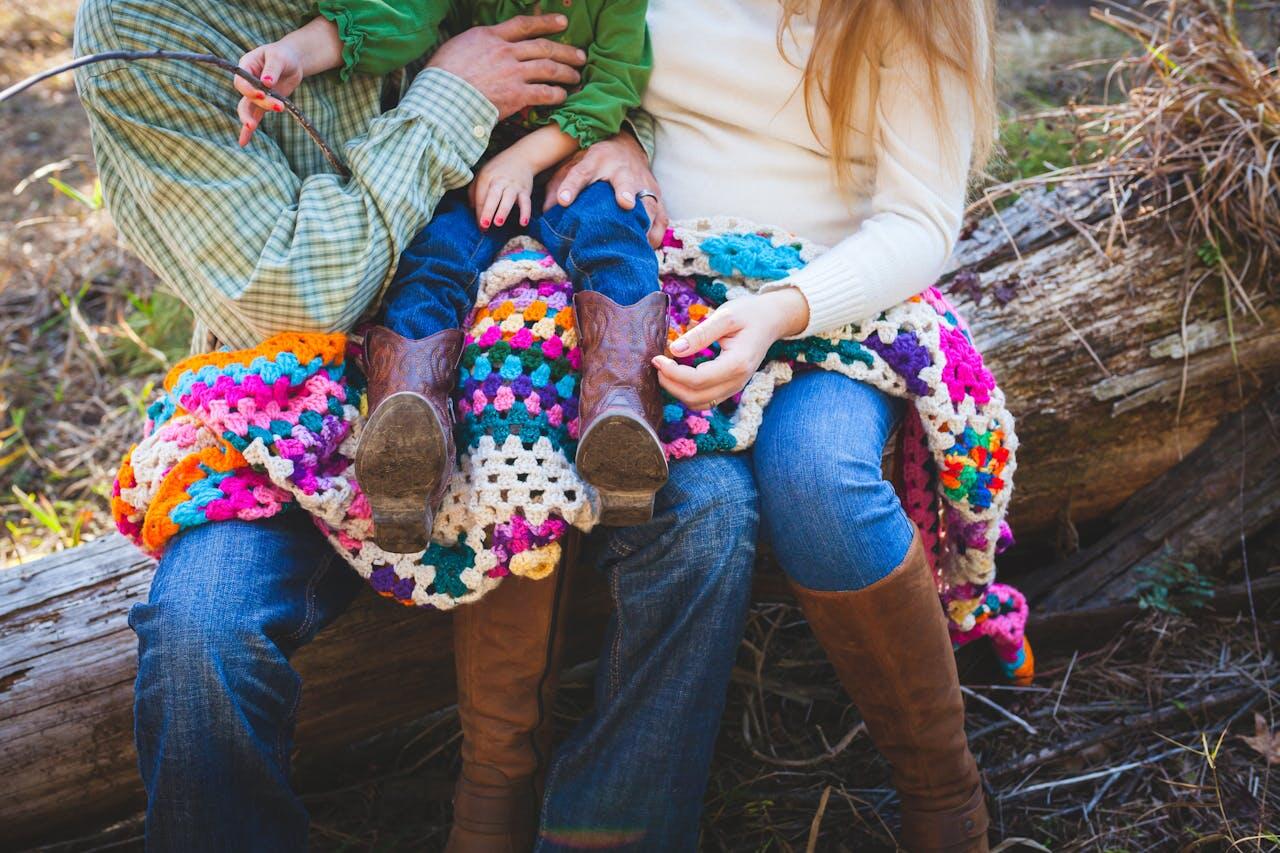 deux parents avec leur jeune enfant assis dehors couverts par une couverture crochetee