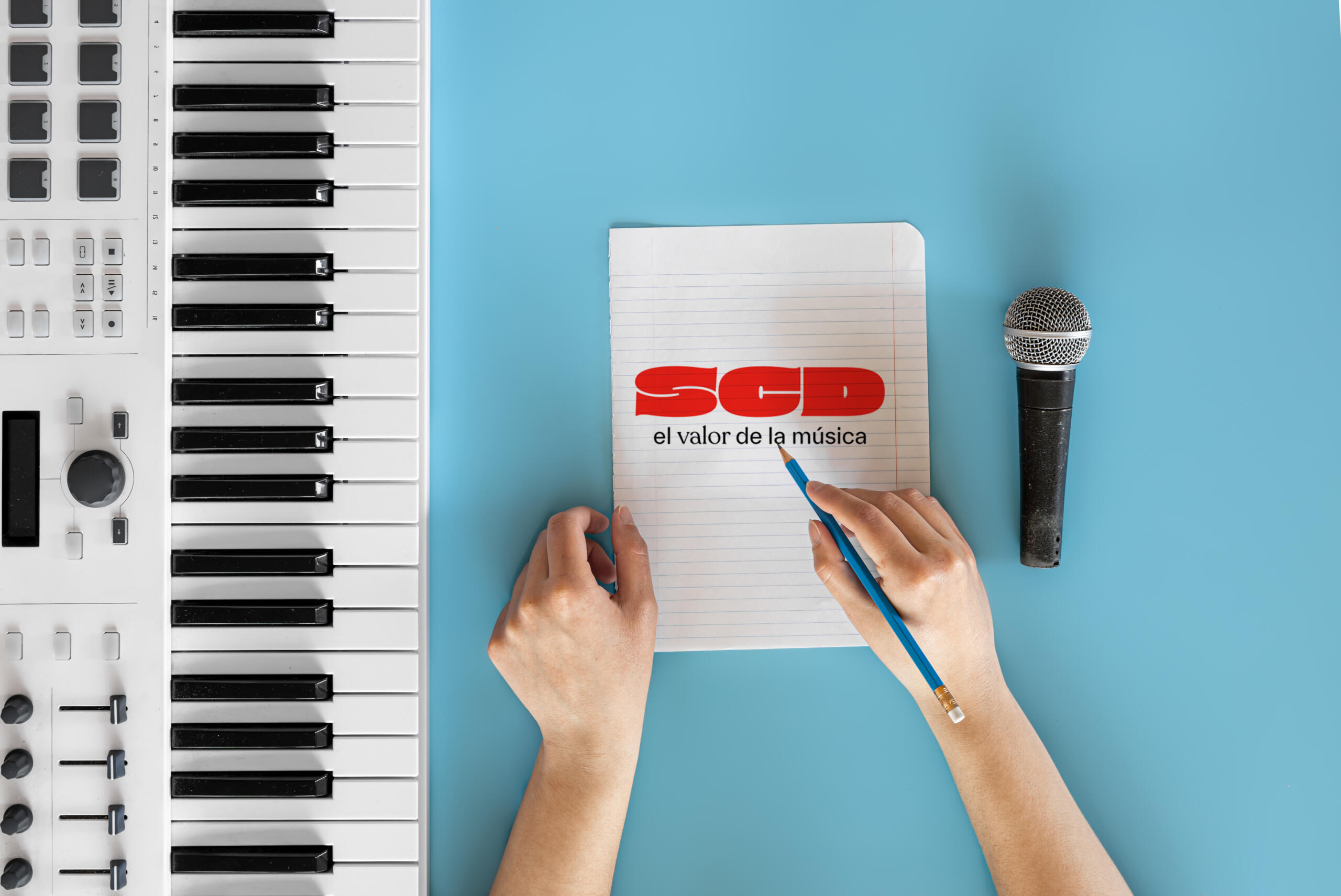 Toma cenital de una libreta con logo de la SCD con una manos sosteniendo un lápiz junto a un micrófono y teclado.