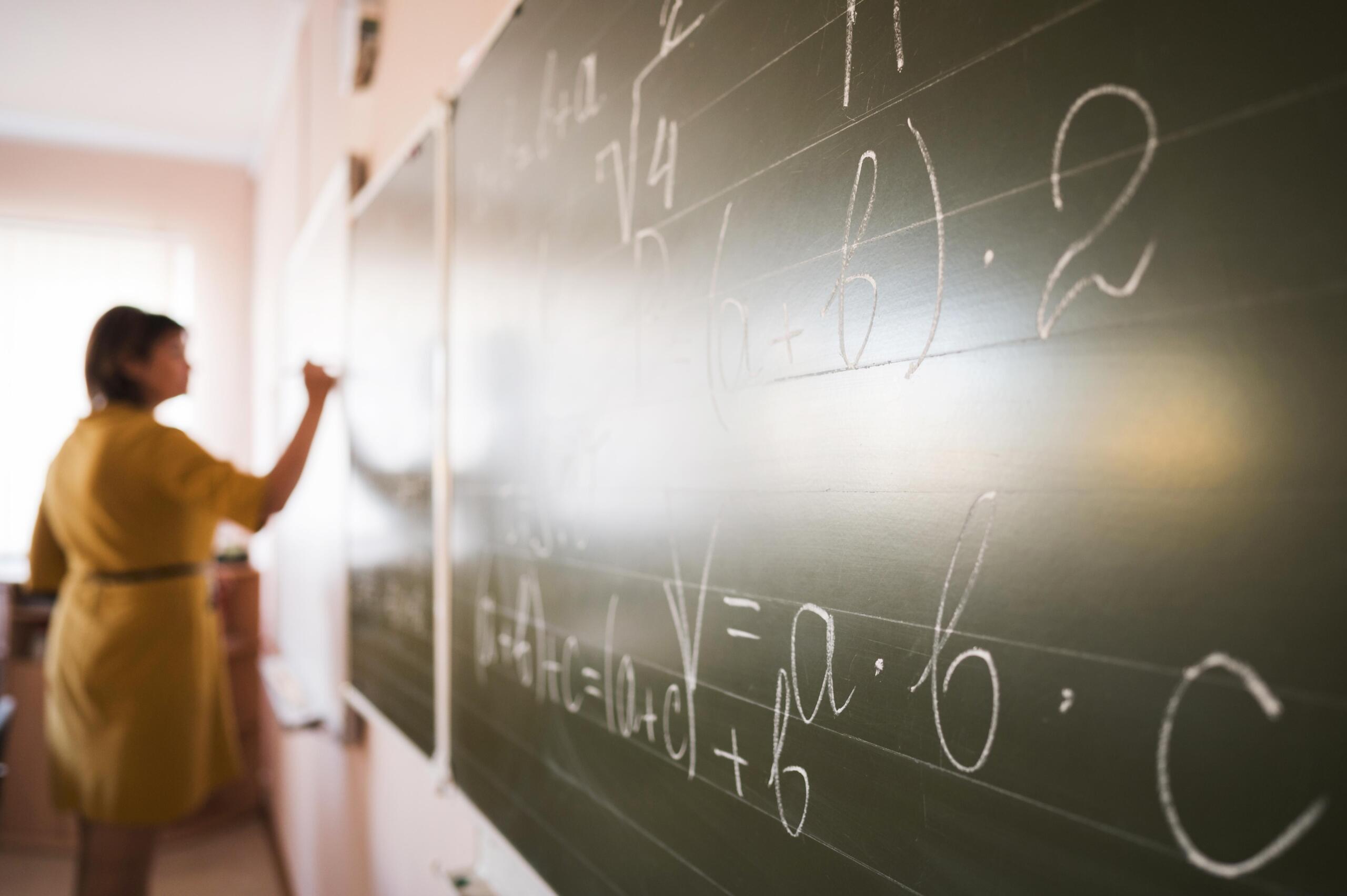 primer plano de una pizarra llena de ejercicios matemáticos y vista de la profesora casi en el fondo escribiendo el otro lado de la pizarra