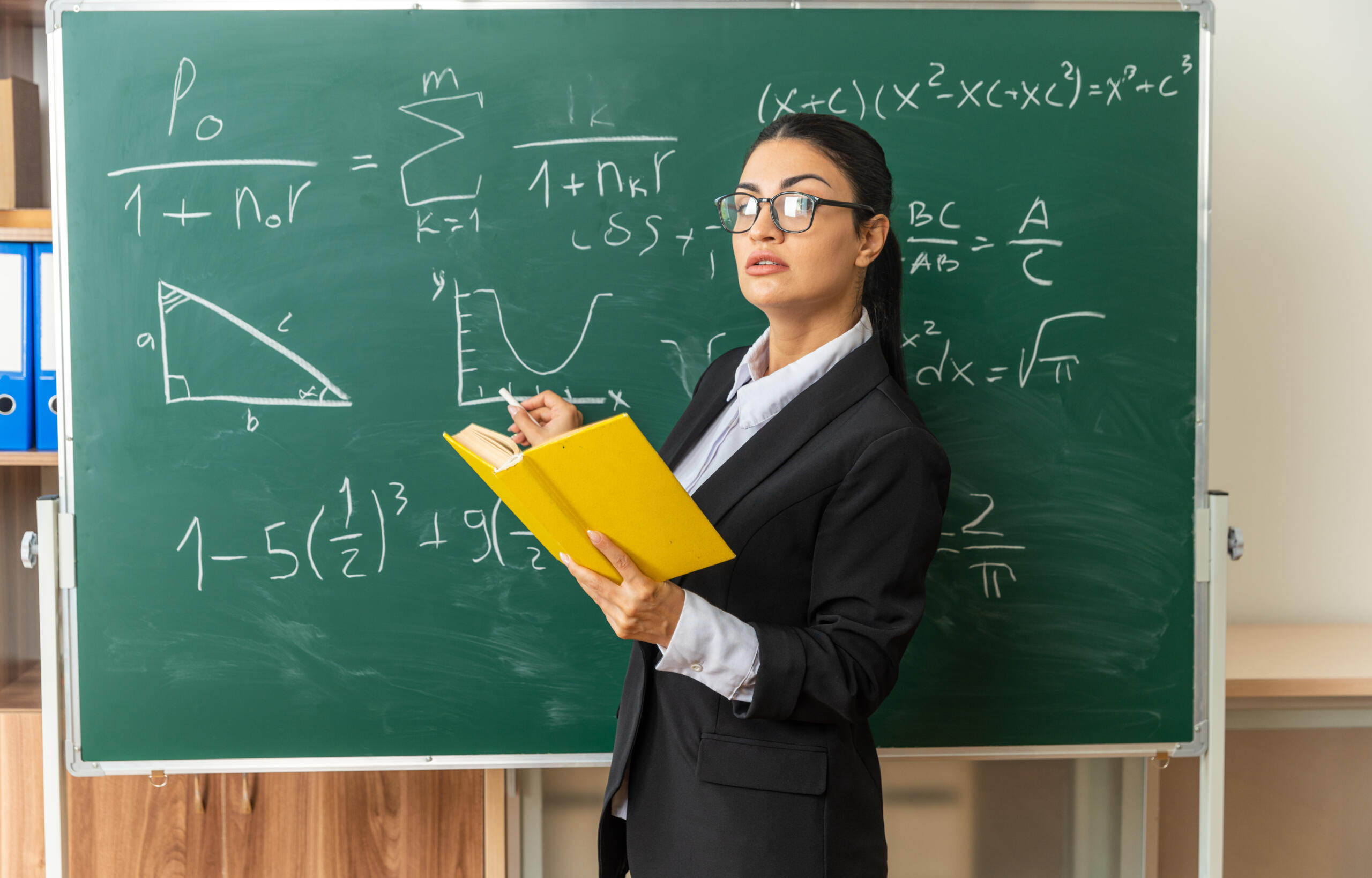 chica con rostro serio y lentes de montura sosteniendo un libro amarillo mientras escribe en una pizarra verde formulas