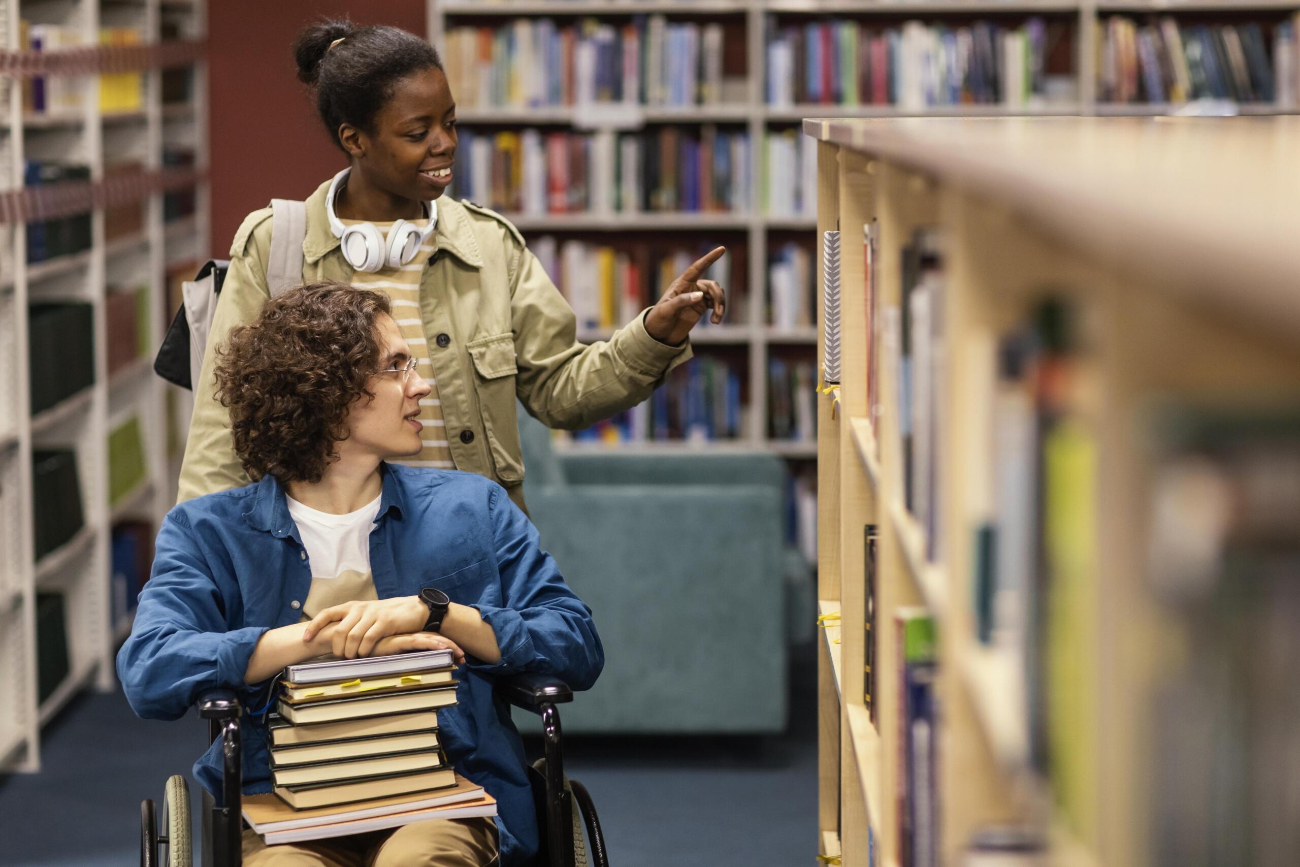chico en silla de ruedas junto a otra estudiante recolectando libros juntos en la biblioteca