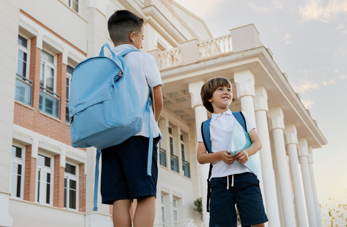 toma contrapicada de dos niños pequeños con sus maletas y uniformes frente a su colegio