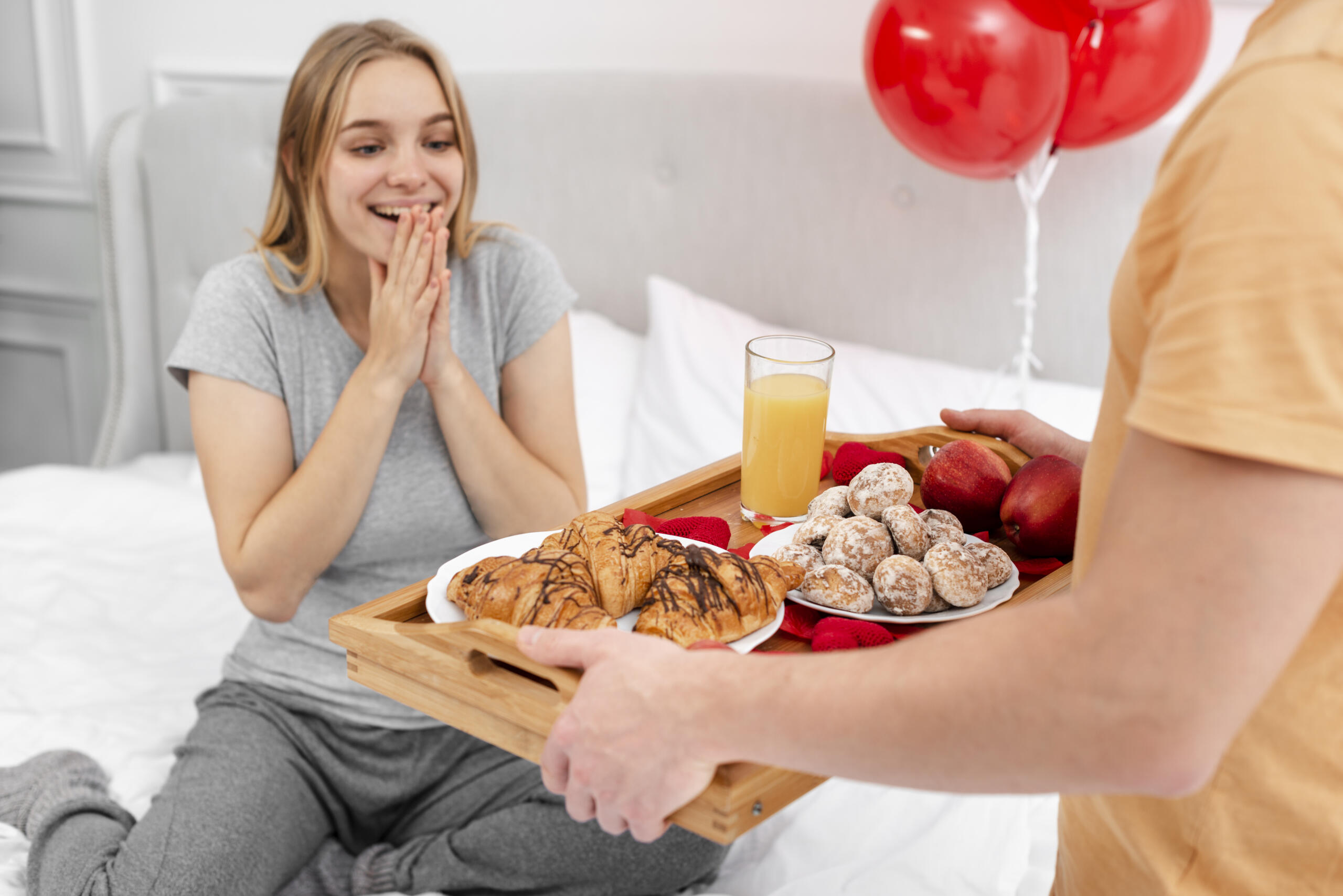 manos de un chico sosteniendo una bandeja con desayuno sorpresa frente a una mujer que está sentada en la cama con rostro alegre y sorprendido