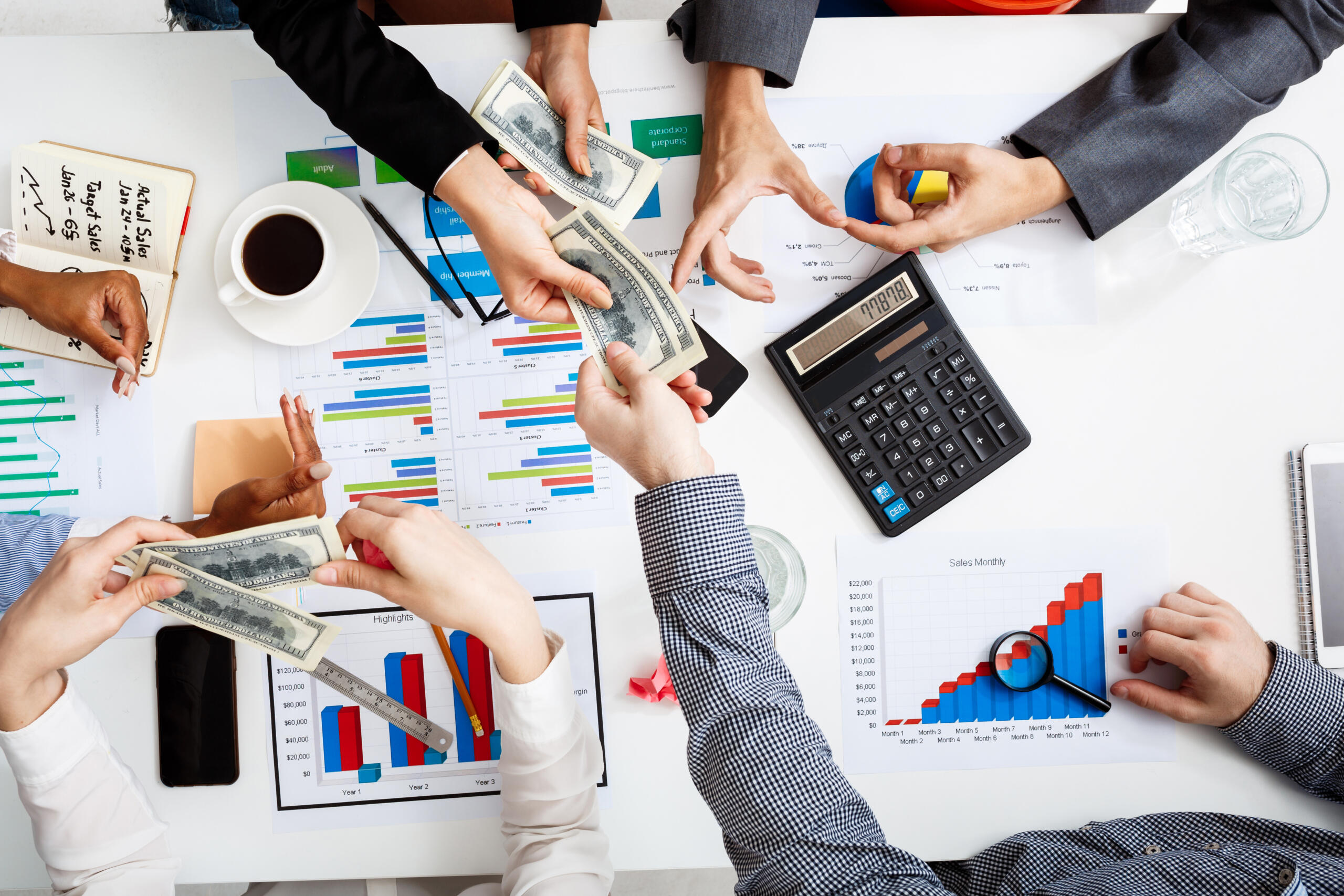 toma cenital de un equipo economista trabajando con estadisticas, data, calculadora y dolares intercambiados sobre la mesa y en manos