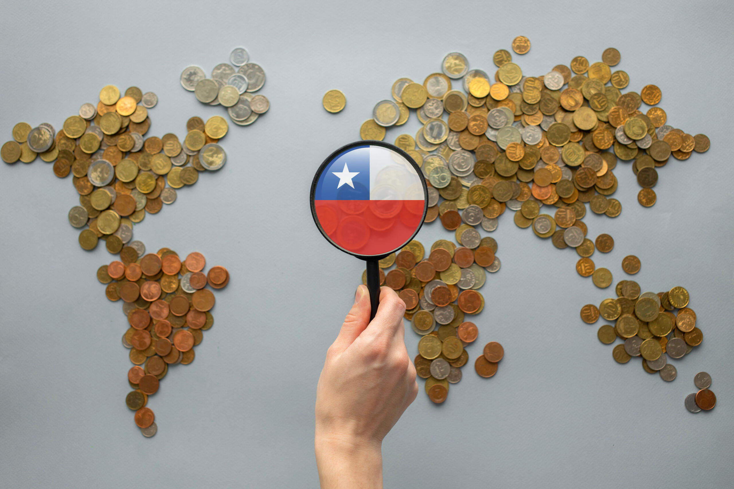 toma cenital de un mapa mundi hecho de moneditas con una mano sosteniendo una lupa con bandera de chile