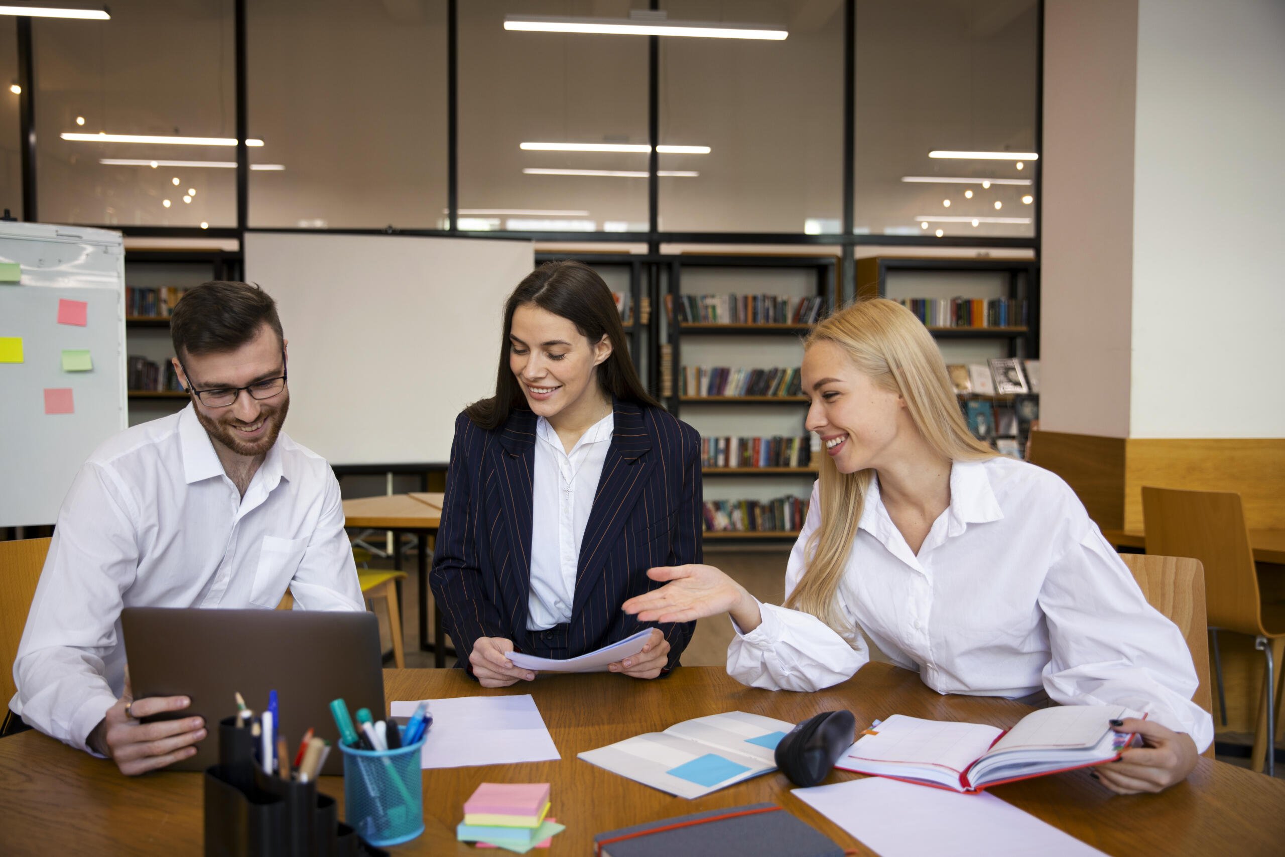 Grupo de tres personas reunidas en una biblioteca revisando apuntes en libros, hojas y una tablet