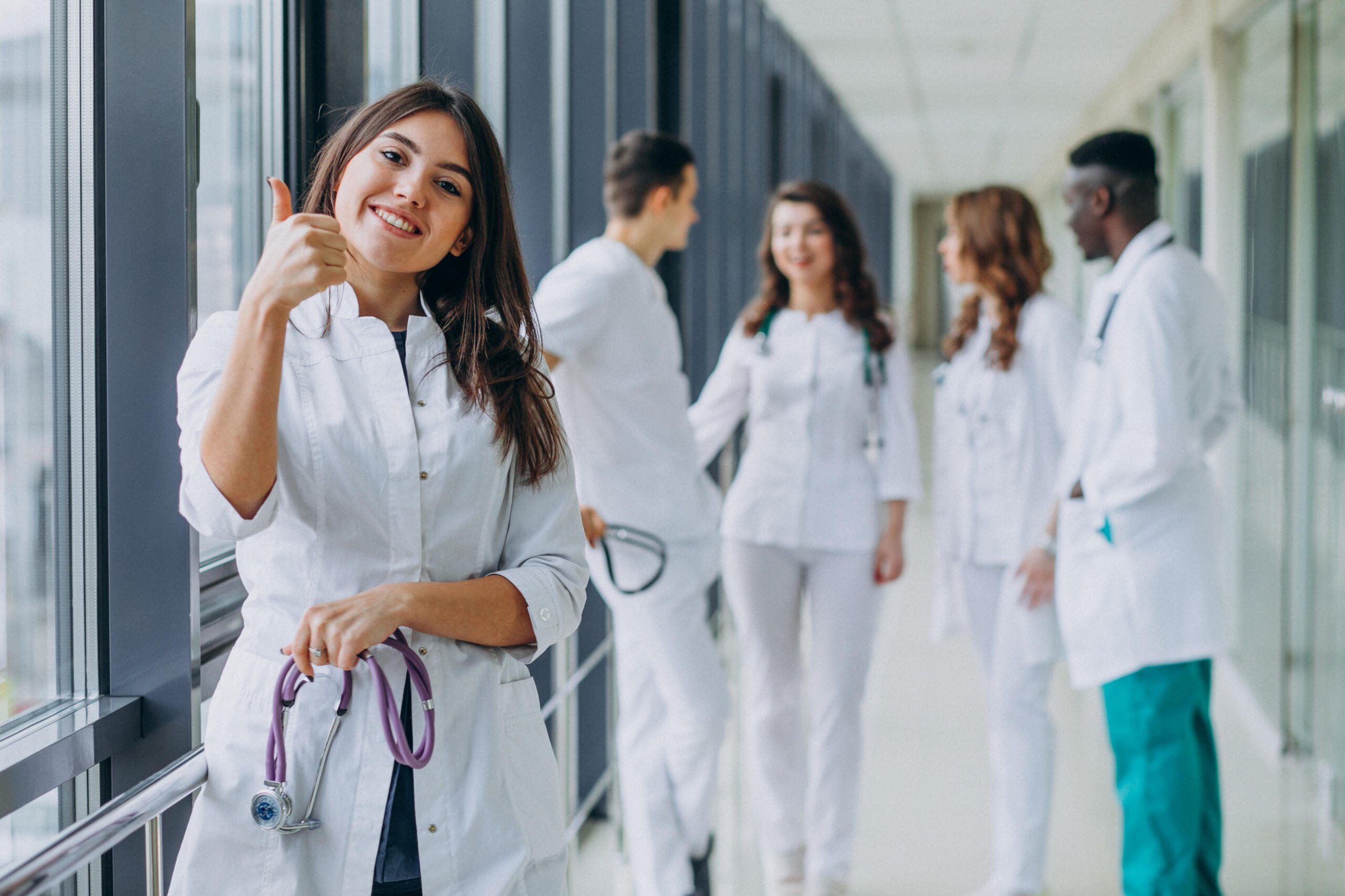 Estudiante de medicina en primer plano feliz levantando un pulgar hacia la cámara y sosteniendo un estetoscopio en medio de los pasillos de un hospital con más médicos y alumnos de fondo.