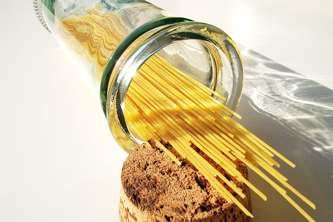 Envase de vidrio con tapa de corcho con pasta tipo espagueti dentro sobre una mesa blanca.