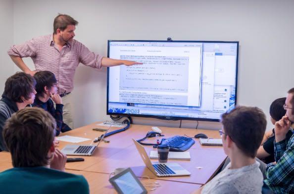 Clase de tecnología usando un televisor e lugar del pizarrón, cada uno de los seis alumnos visibles en la imagen con su computadora.