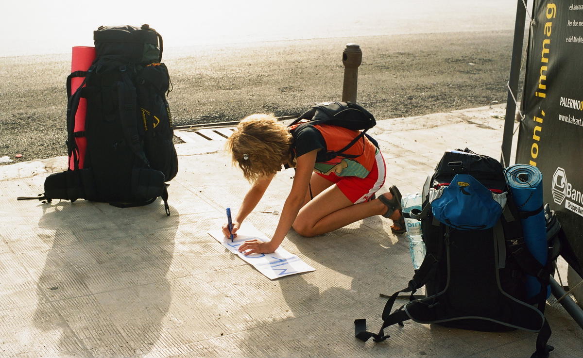 Chica arrodillada en un mirador marcando en el mapa su trayecto, con 2 mochilas viajeras junto a ella.
