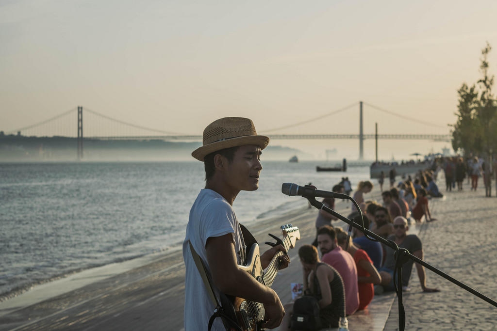 Chico con sombrero tocando guitarra y cantando en una calle concurrida junto al mar