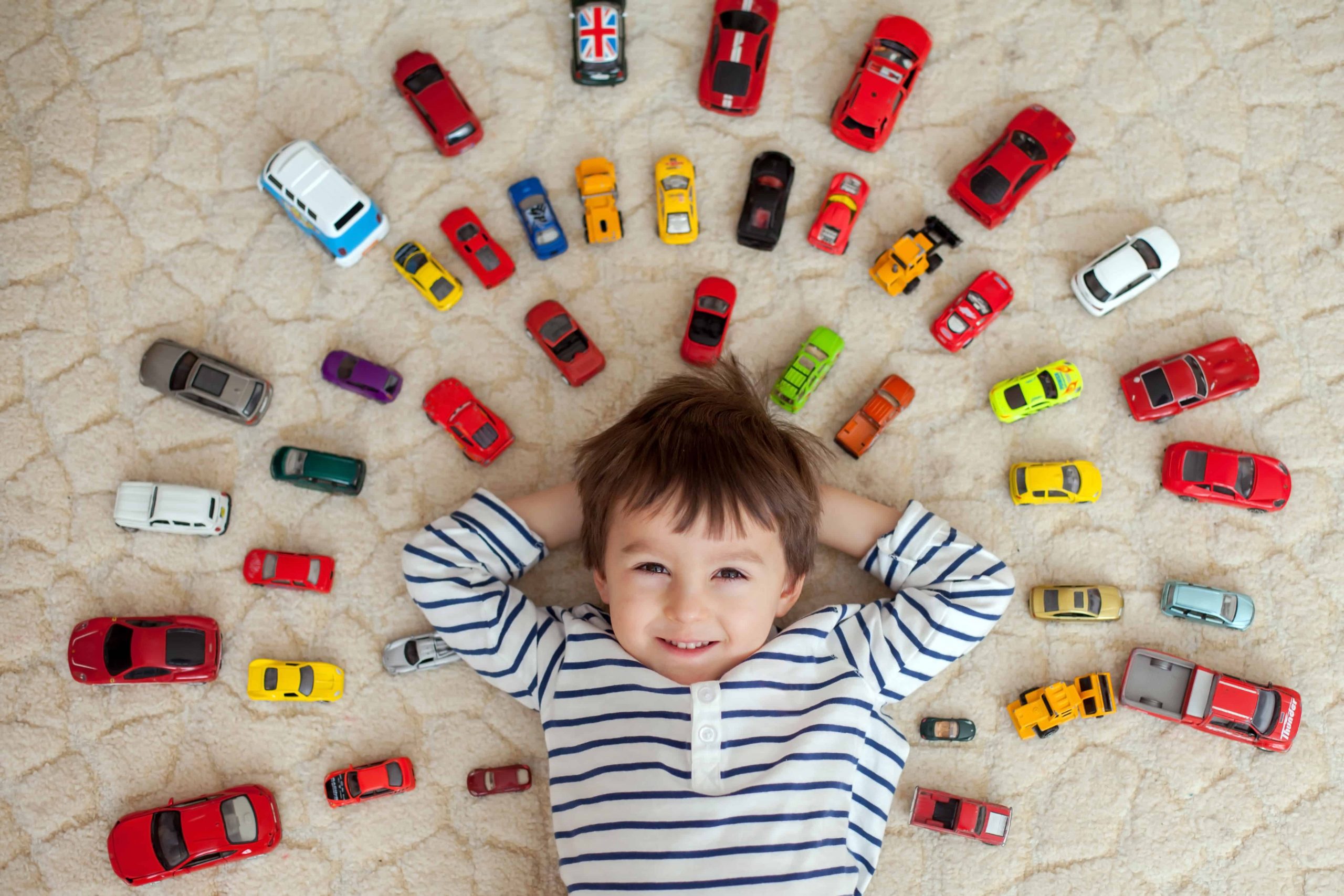 Niños recostado de para atrás en el piso, con sus carros alineados a su alrededor.