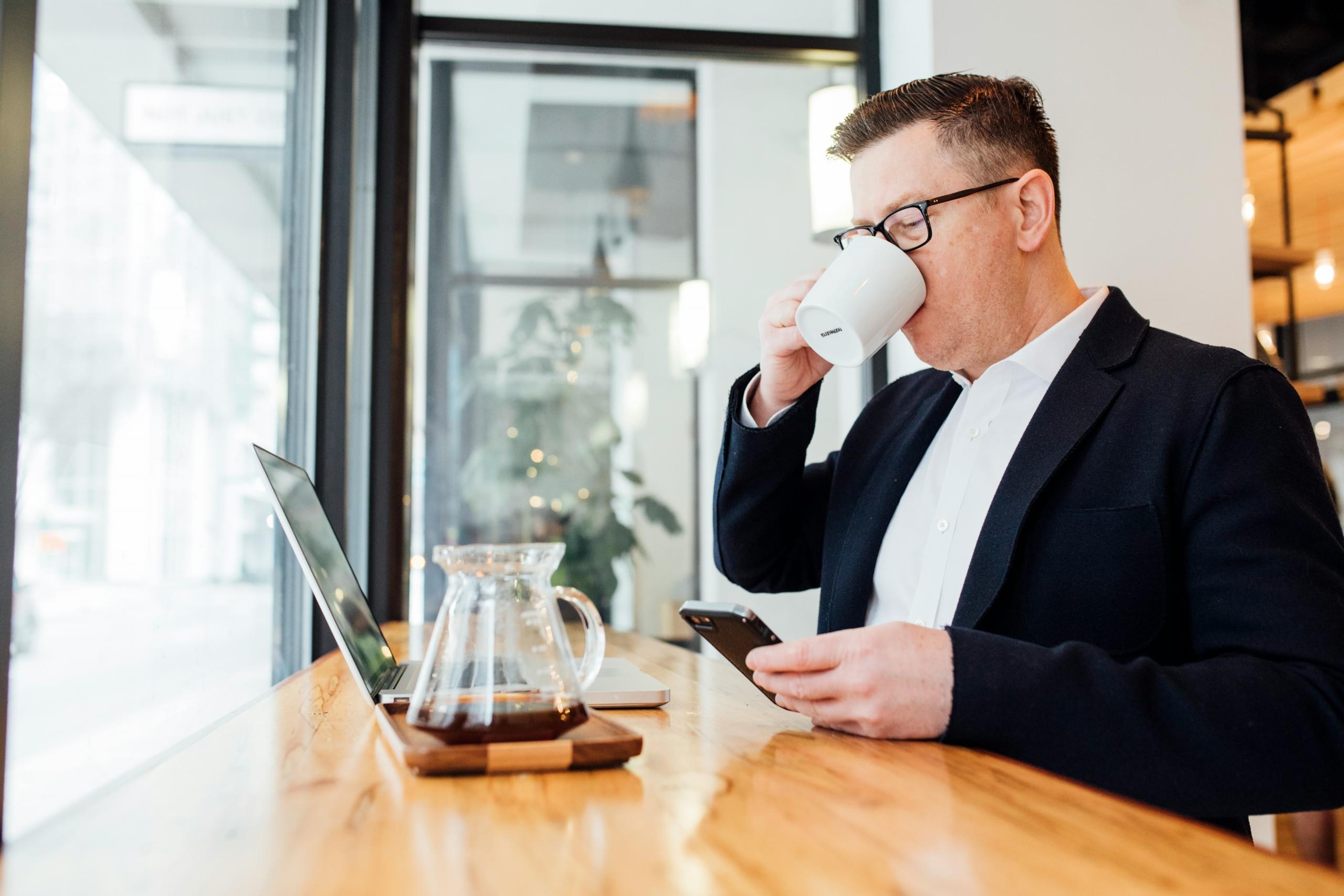 Hombre sentado bebiendo una taza de café mientras revisa su telefono y computadora al tiempo