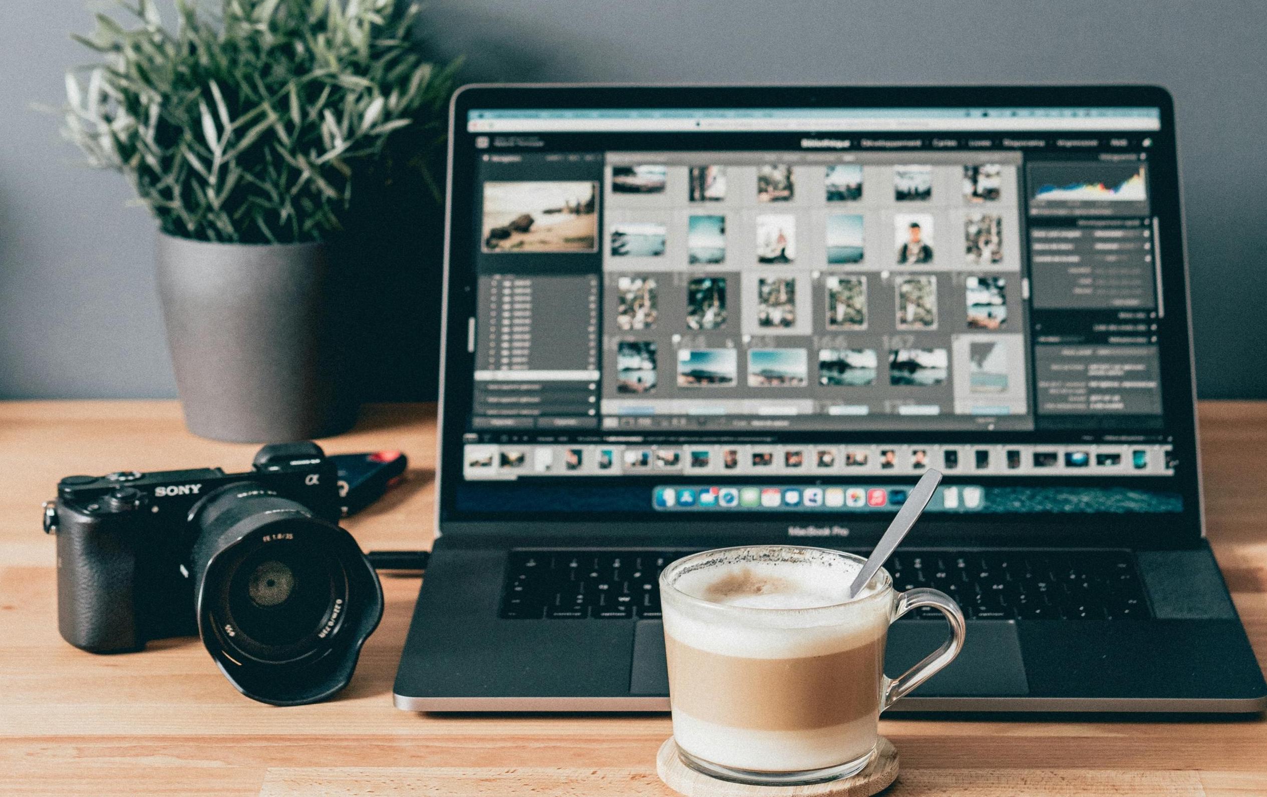 Imagen que muestra un laptop con un programa de edición de fotografias, una cámara digital y una taza de café sobre una mesa.