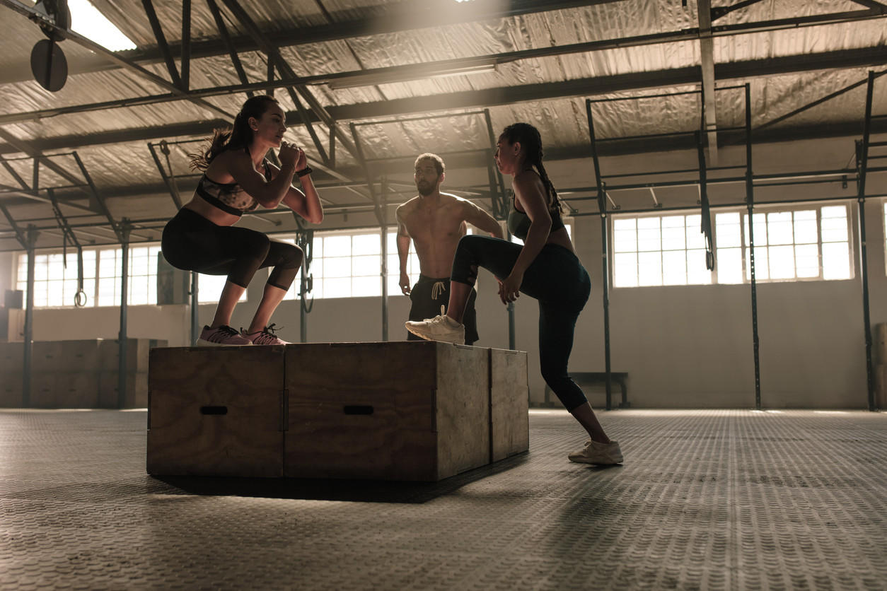 Dos chicas y un entrenador realizando el ejercicio de salto de cajón, en una sala grande.
