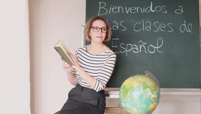 Profesora sentada delante de un tablero con un libro en la mano y un mapamundi junto a ella.