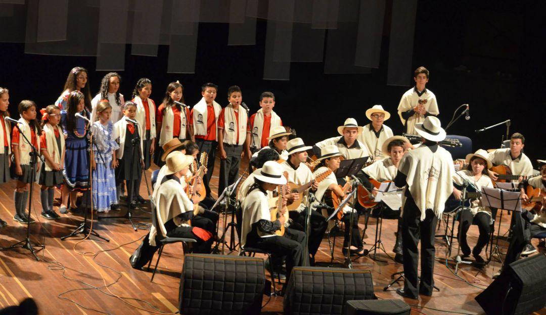 Gran orquesta con traje tradicional de región andina interpretando bambucos. Atrás un coro de niños cantando