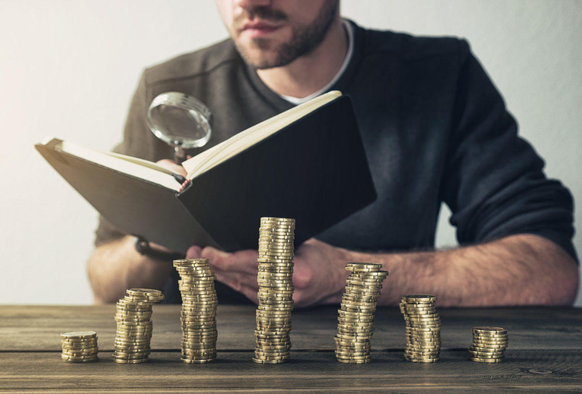 Hombre estudiando un libro con una lupa, frente a una barra de graficas hecha con monedas.