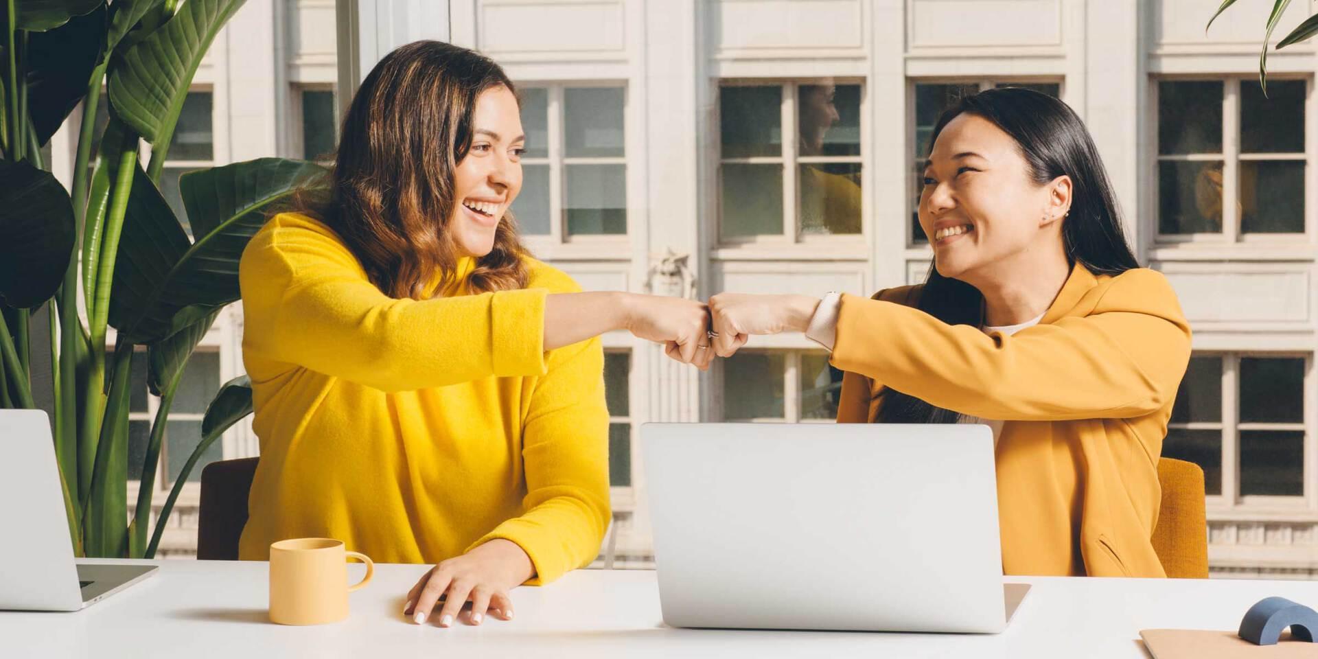 Amigas sentadas frente a un escritorio, uniendo sus puños y riendo.