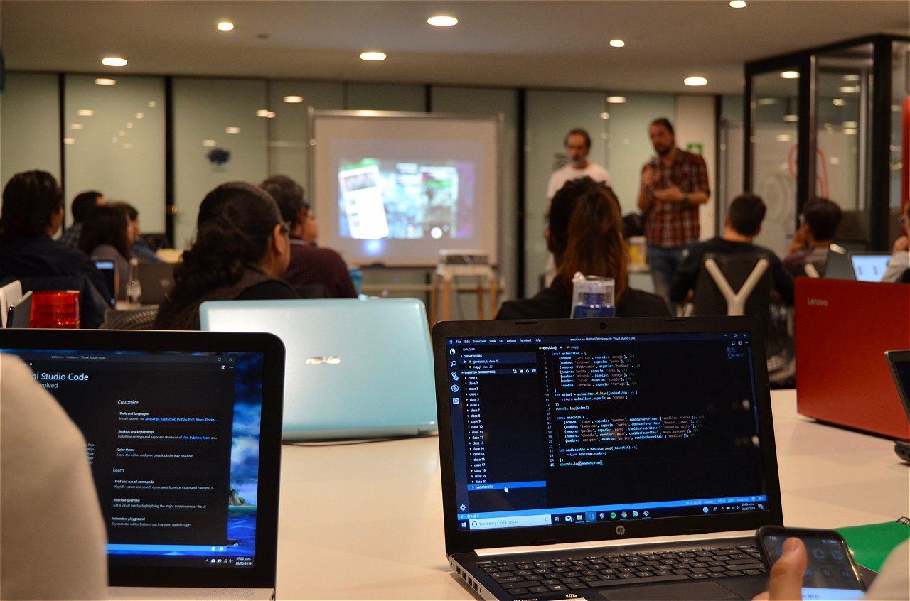Computadoras mostrando un código. De fondo estudiantes frente a profesores de programación.