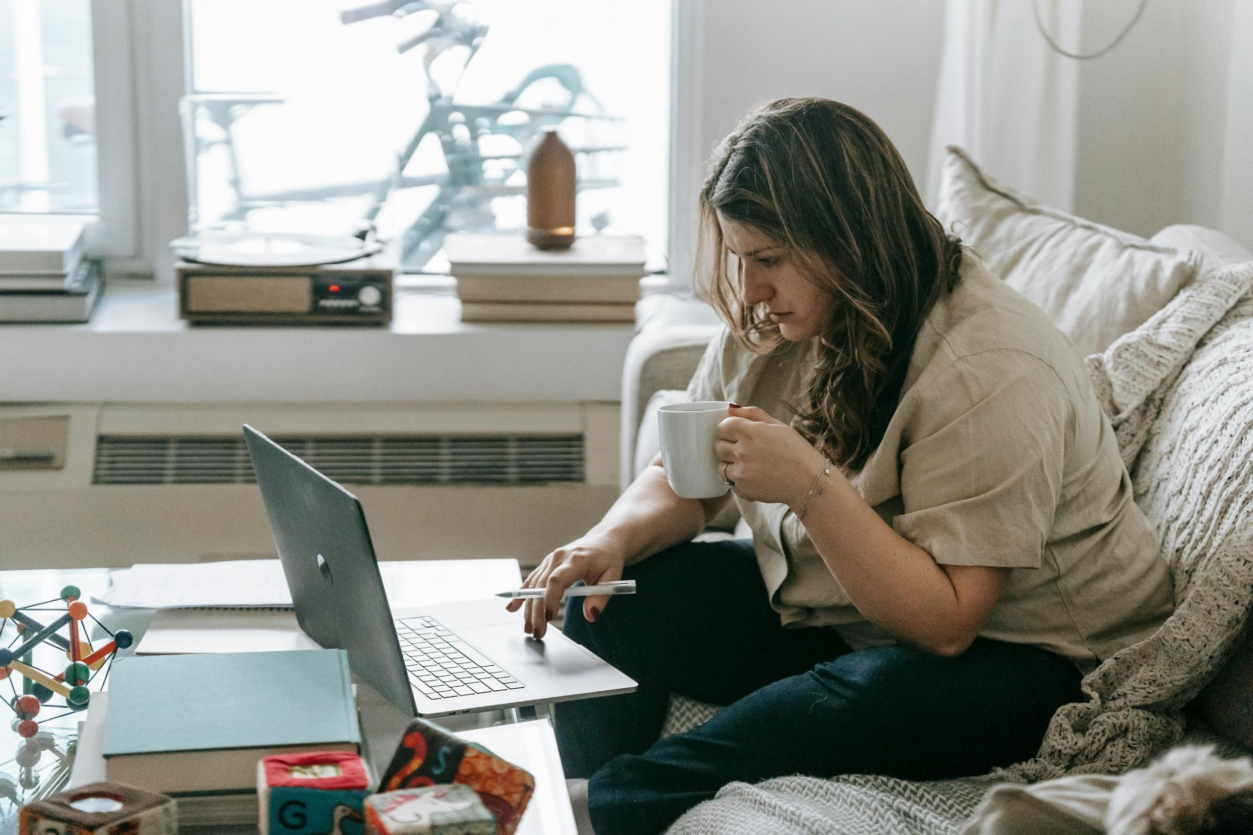 Mujer estudiando online a través de un computador, en la mesa se observan algunos libros abiertos y otros objetos.