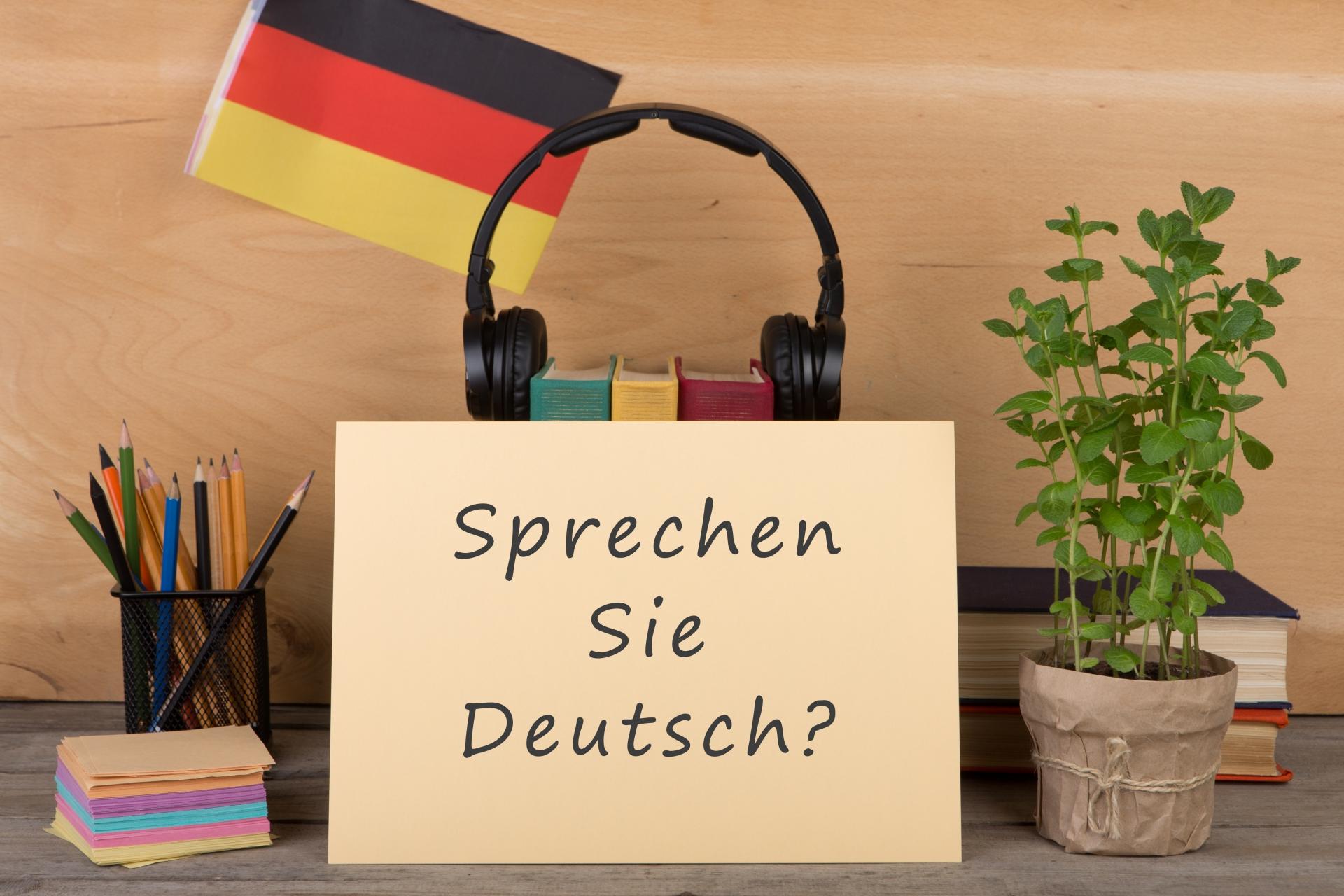 Escritorio con lápices, libros, audífonos y una bandera de Alemania. Al frente una hoja que dice: Sprechen sie Deutsch
