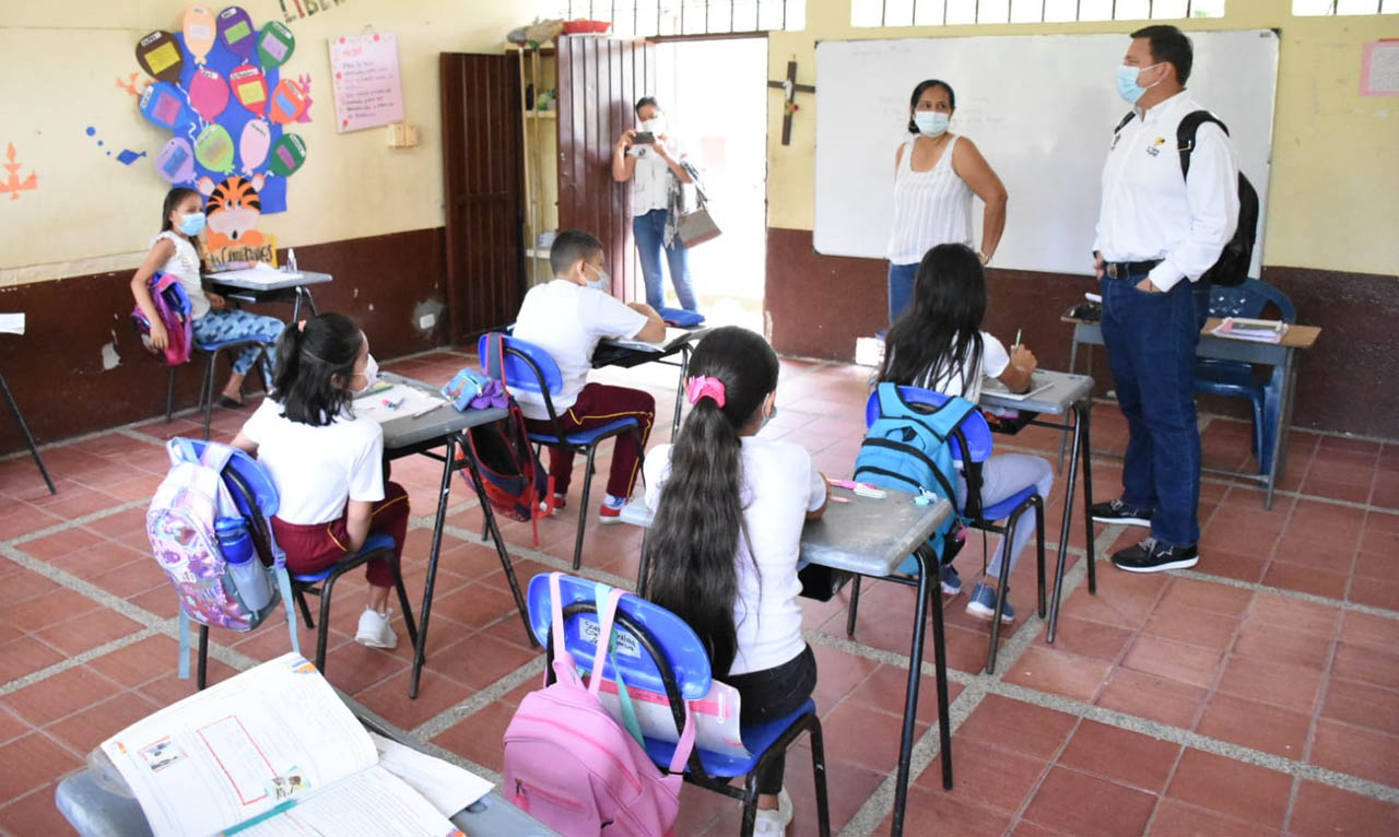 Niños sentados en un salón de clases, y frente a ellos una profesora y funcionarios del gobierno.