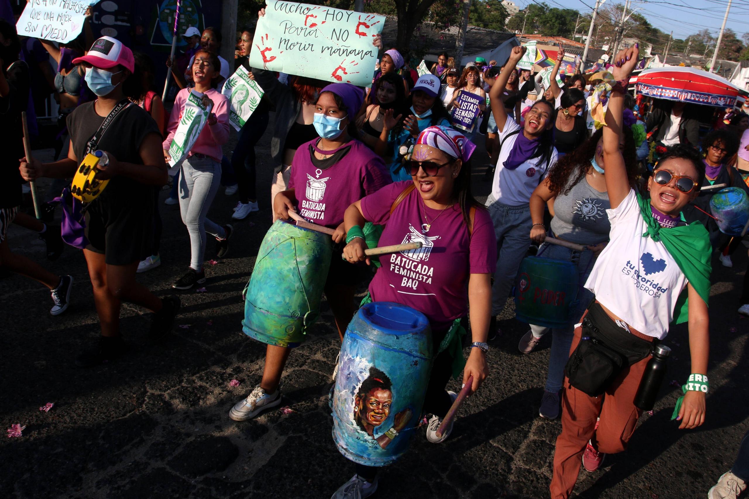 Mujeres marchando por las calles con tamboras y camisetas moradas.