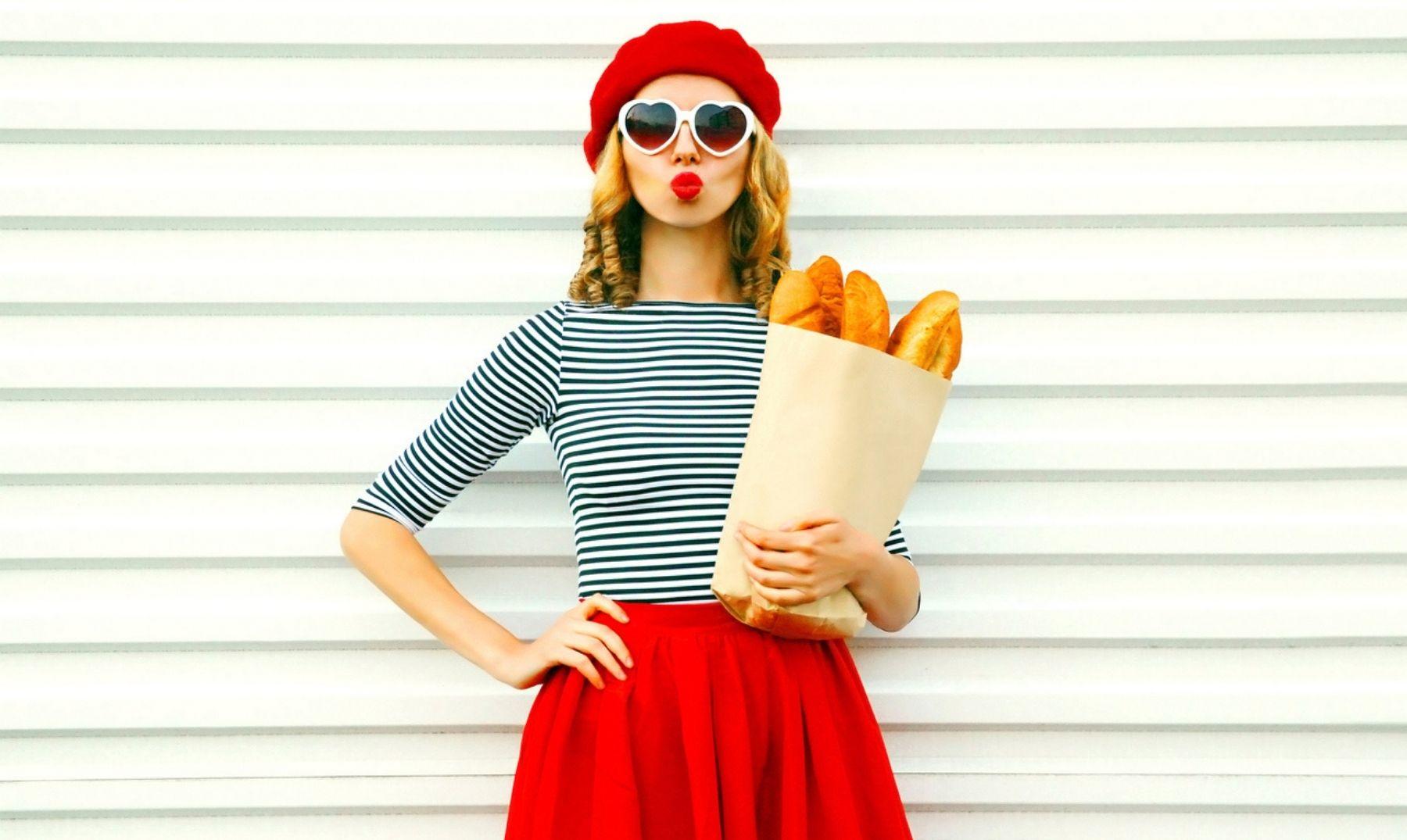 Chica vestida con una falda roja, buso a rayas, gafas con forma de corazón y boina, sosteniendo una bolsa de papel llena de panes.
