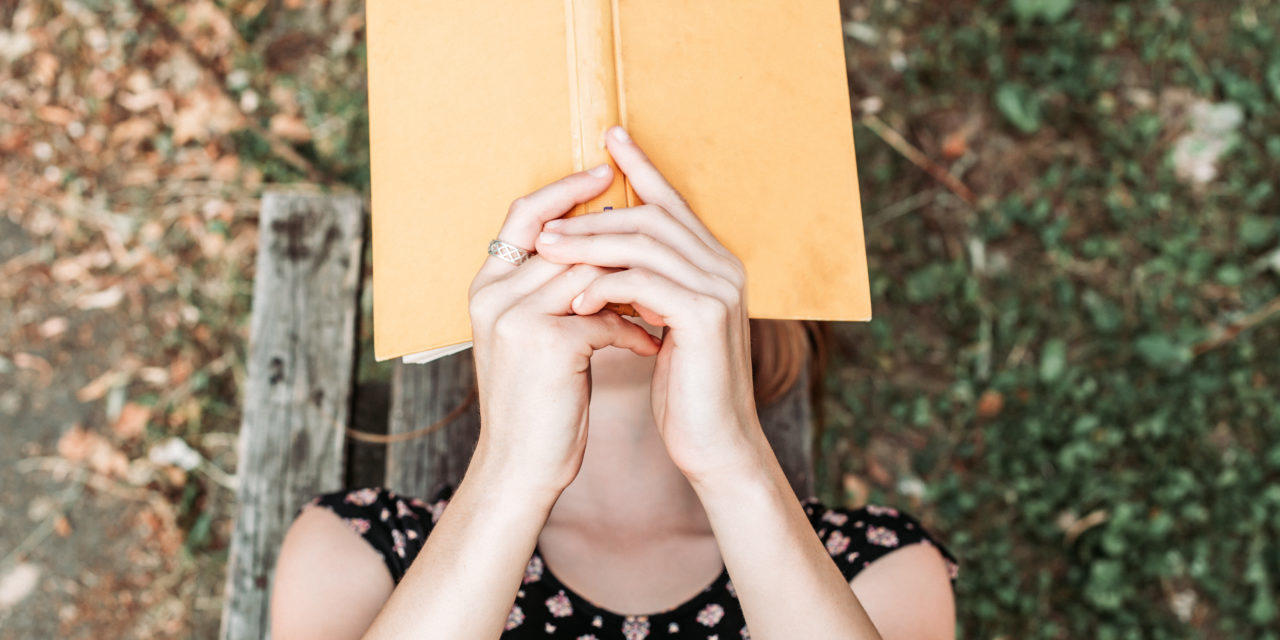 Chica recostada en la hierba con un libro cubriendo su rostro.