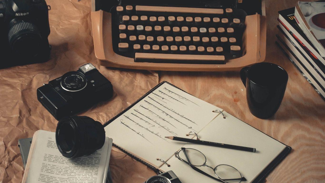 Máquina de escribir junto a un libro abierto, unas gafas y un lápiz.
