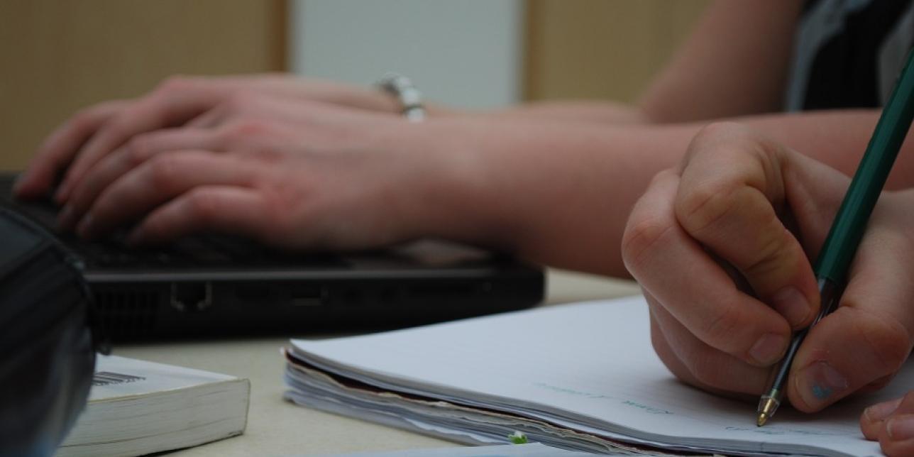 Manos escribiendo en un cuaderno, junto a otras manos que están escribiendo en una computadora.