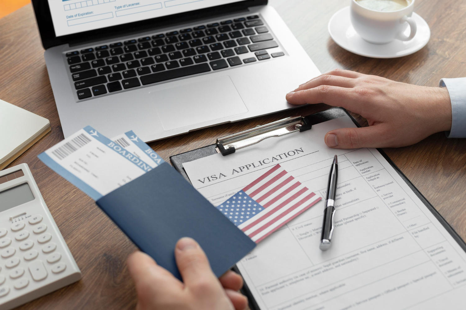 Persona frente a una laptop sosteniendo el pasaporte con unos tiquetes y sobre el escritorio un formulario de aplicación a visa