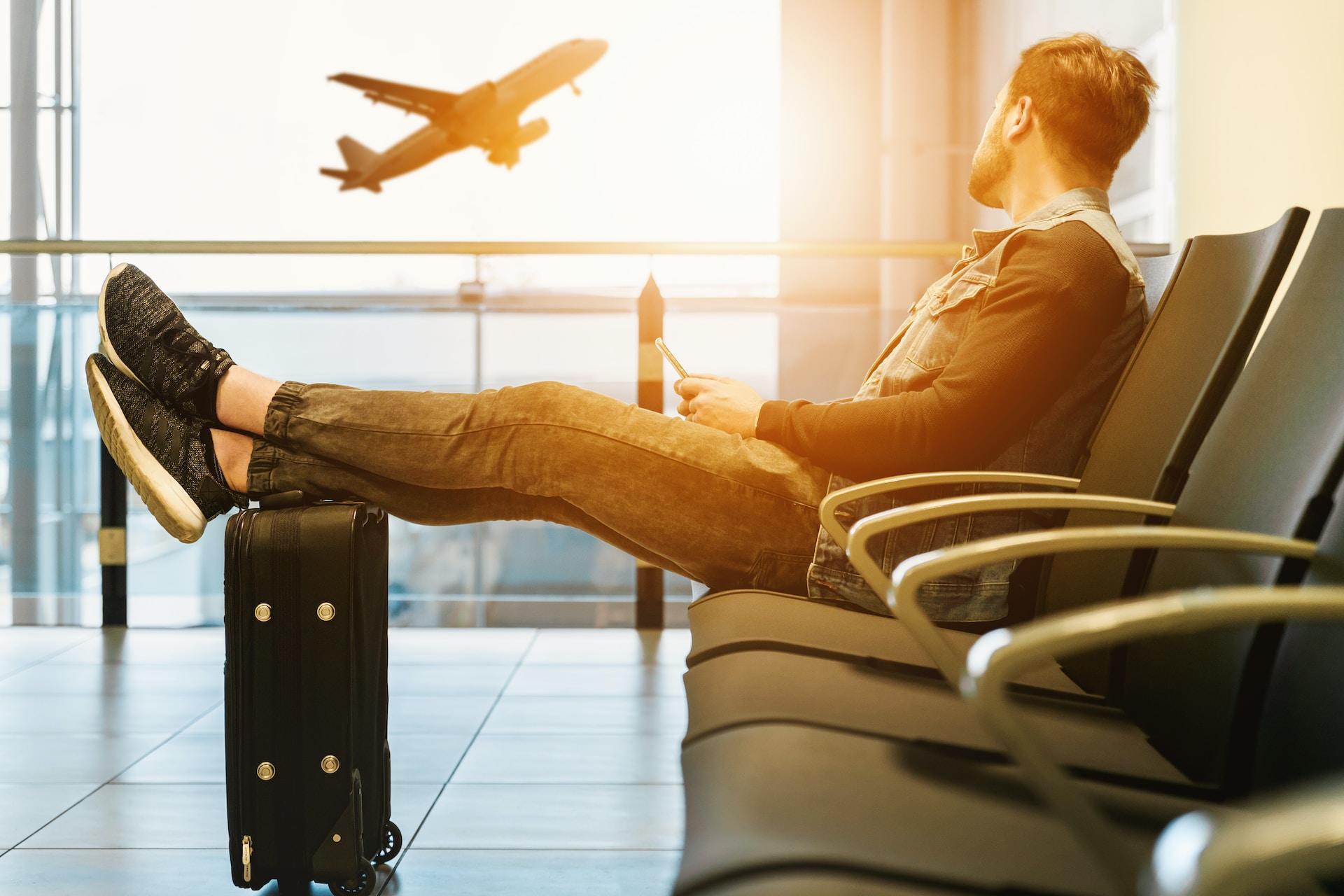 Hombre en sala de espera de aeropuerto, mira un avión despegando por la ventana