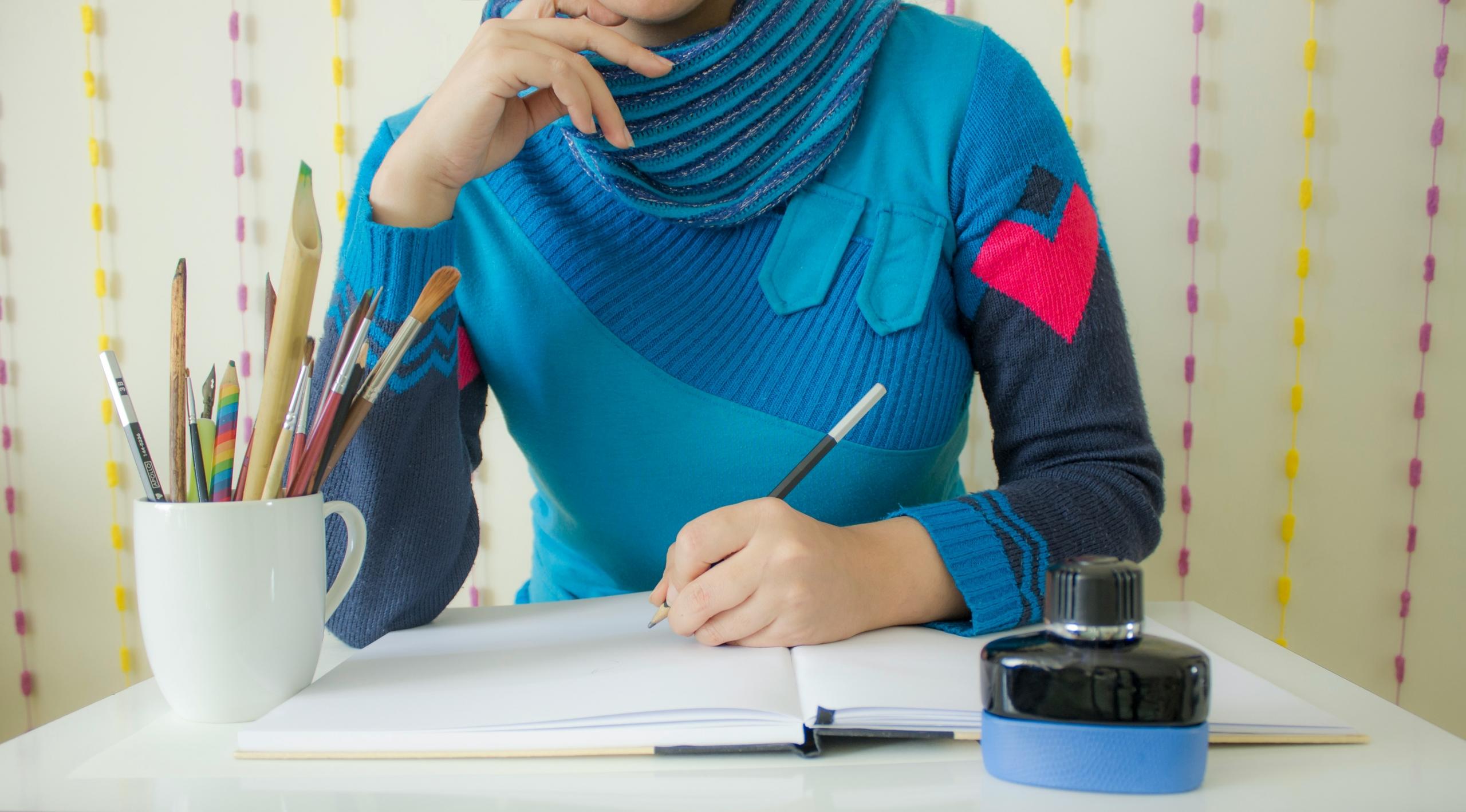 La imagen muestra a un estudiante enfocado en su escritura, con una gama de útiles escolares como lápices y pinceles dispuestos en una taza, y un tintero al lado, lo que sugiere la preparación para una sesión de estudio o arte