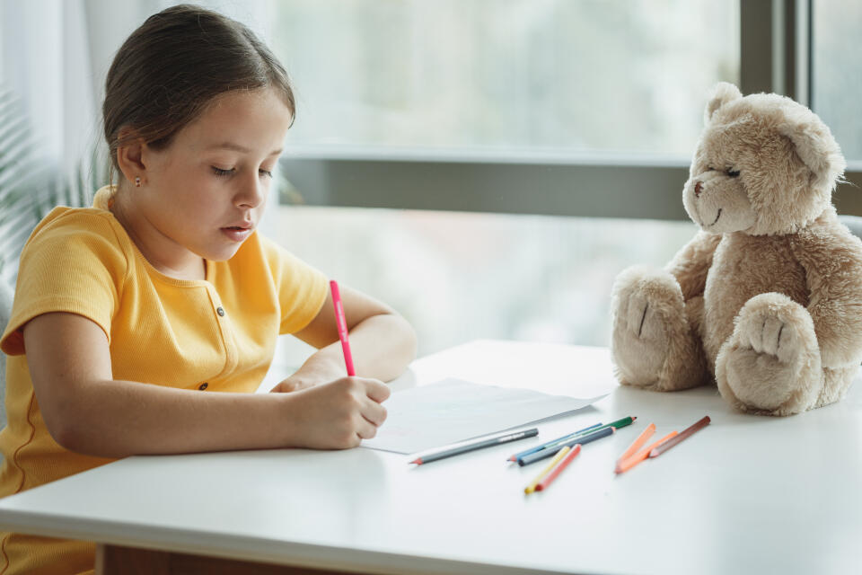 Niña estudiando en casa, escribe con lápices de colores y tiene a su osos Ted al lado.