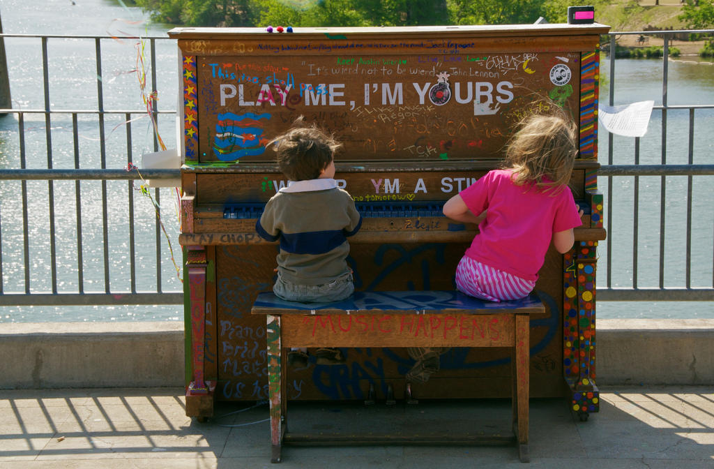 Dos niños tocando un piano acustico colocado a la interperie sobre un paso peatonal. El piano tiene varios textos y mensajes de diferentes colores y aparenta una pared llena de grafitis entre el cual destaca la frase en inglés: "Tocame, Soy tuyo".