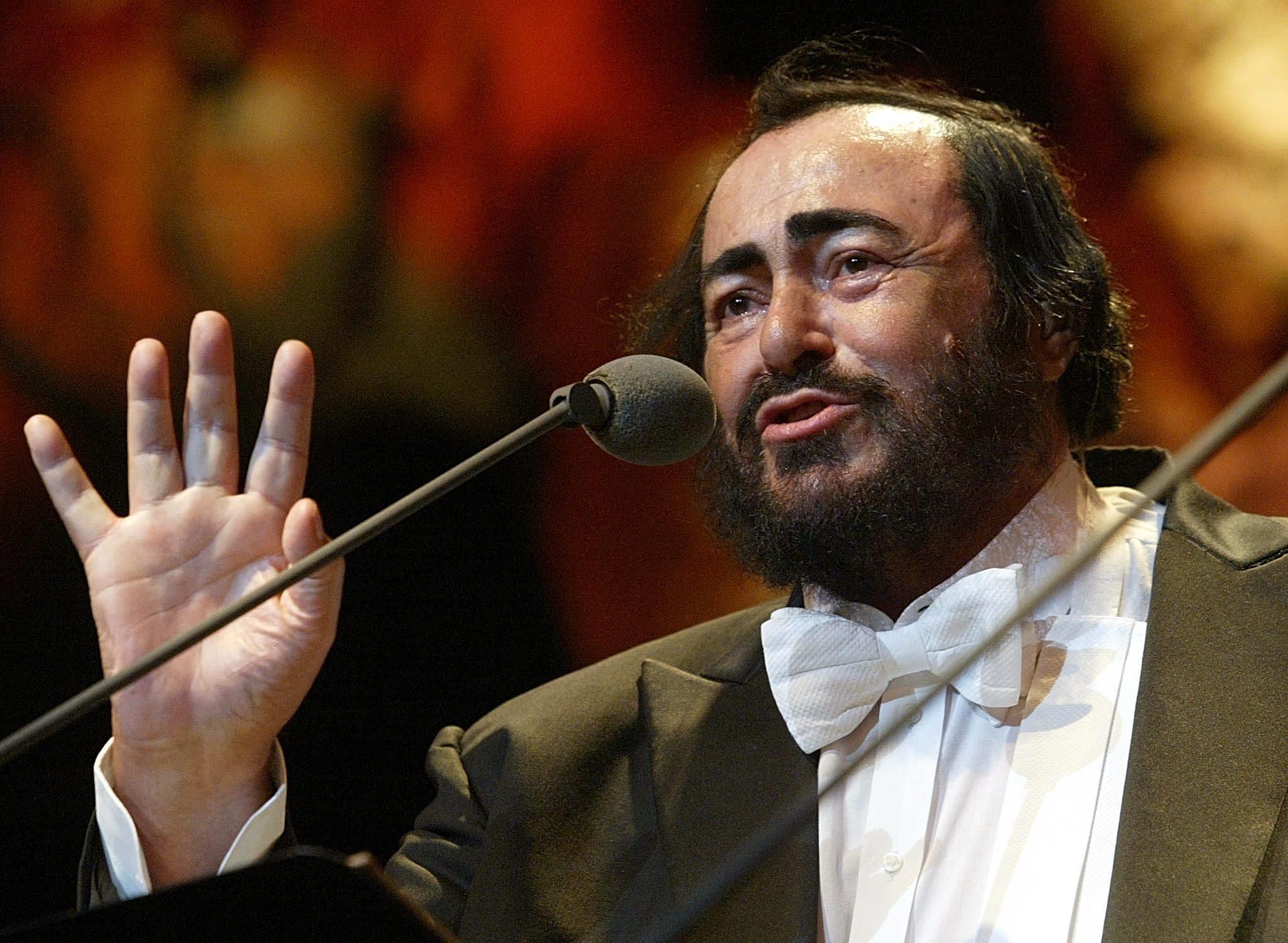 Pavarotti con la mano abierta hacia arriba frente a un micrófono