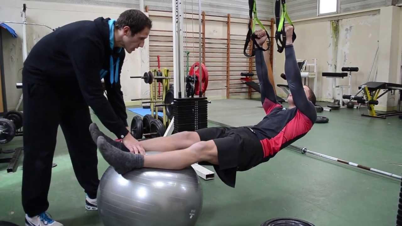 Hombre en el gimnasio haciendo abdominales apoyado en un Softball y con ayuda del entrenador