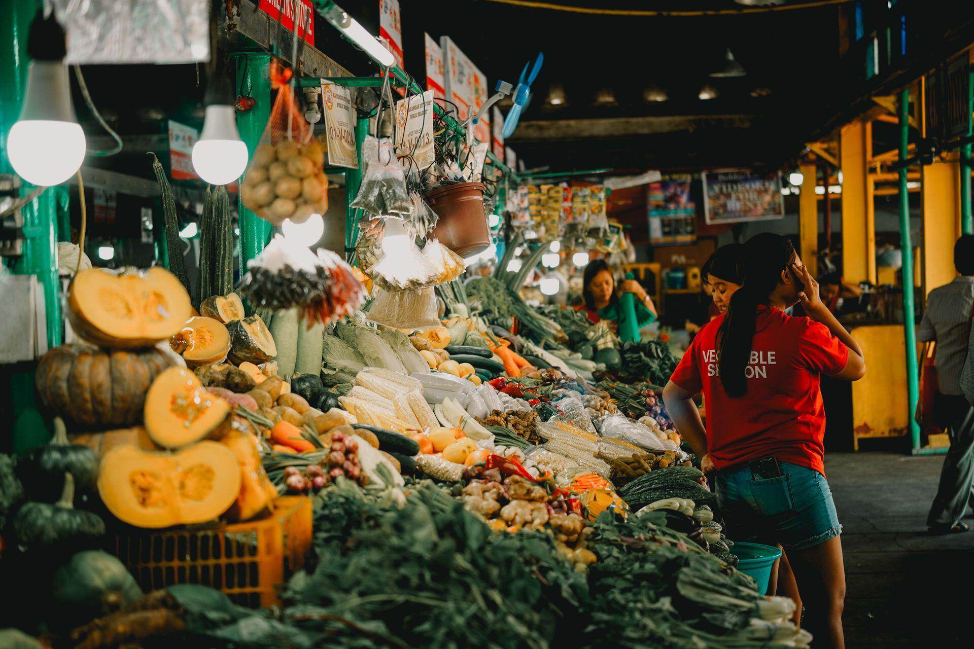 Un mercado cubierto que exhibe una variedad de frutas y verduras frescas, con gente haciendo sus compras.