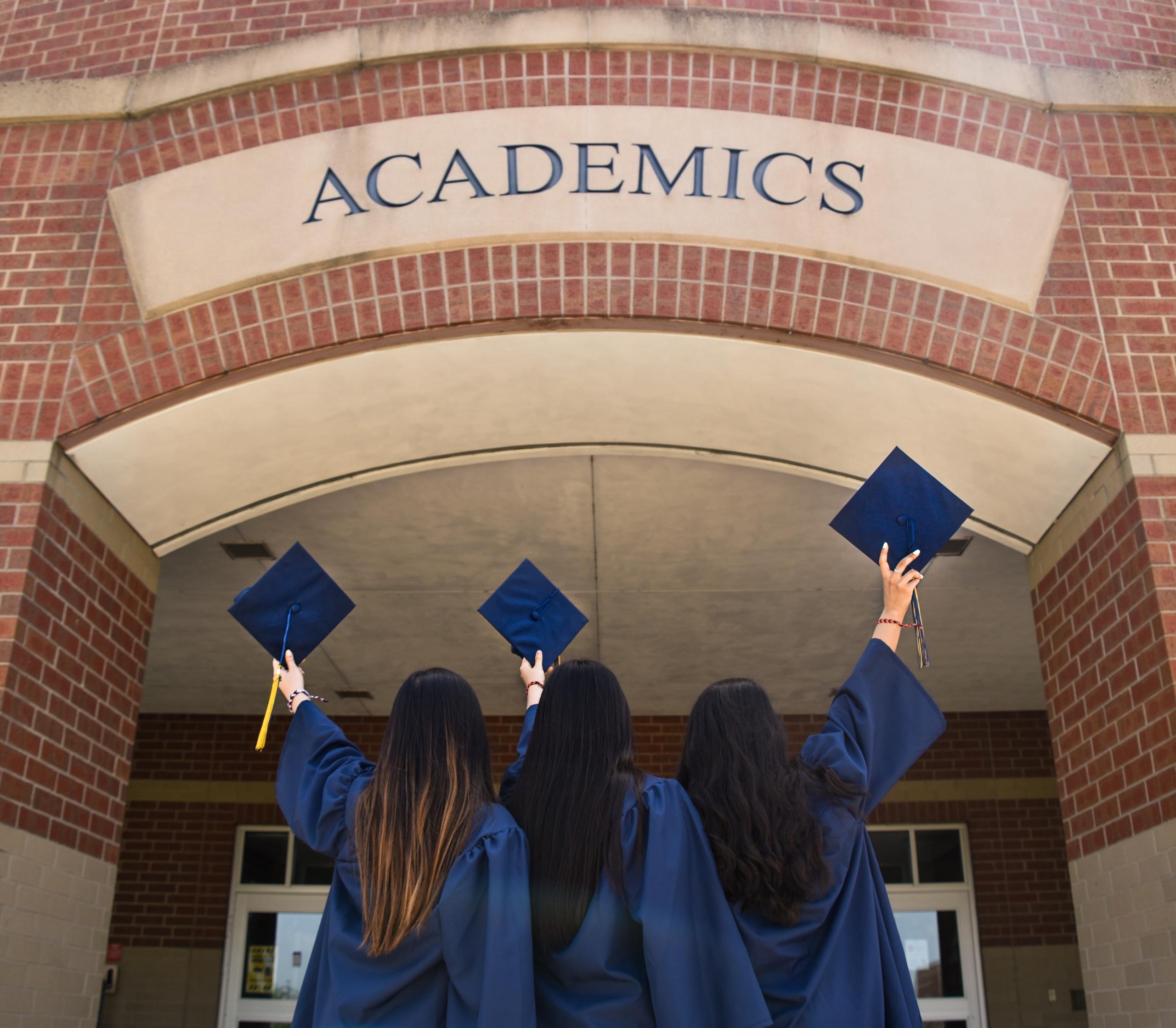 3 chicas alzando su birrete frente aun a puerta que tiene la palabra "ACADEMICS"