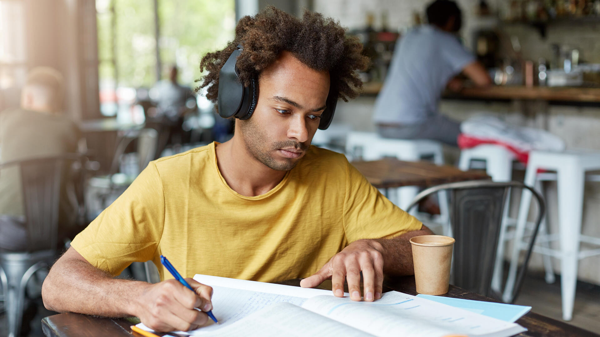 Joven universitario concentrado, con auriculares y apuntes, estudiando en una cafetería.