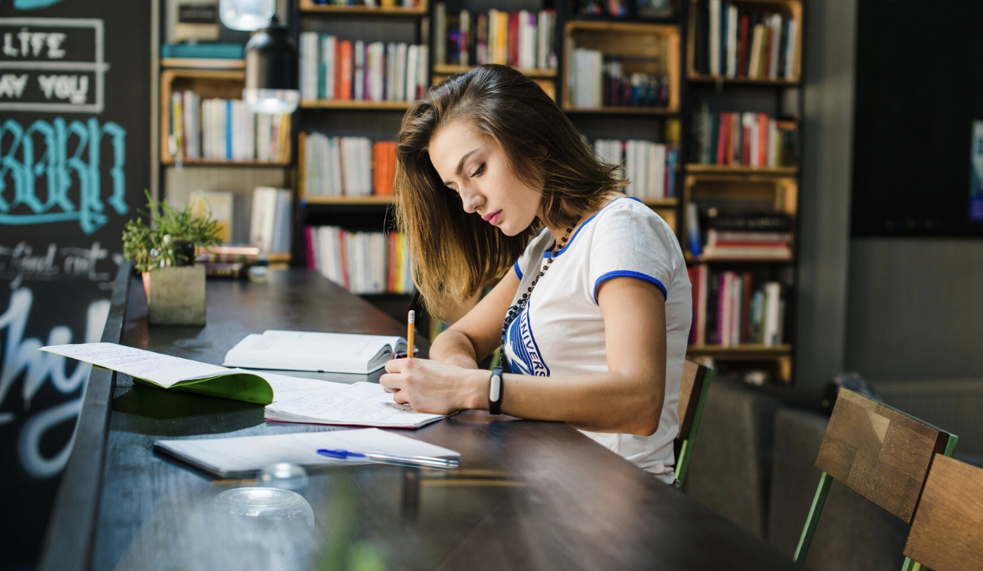 chica de perfil escribiendo con sus libros abiertos con una mesa en medio de la biblioteca