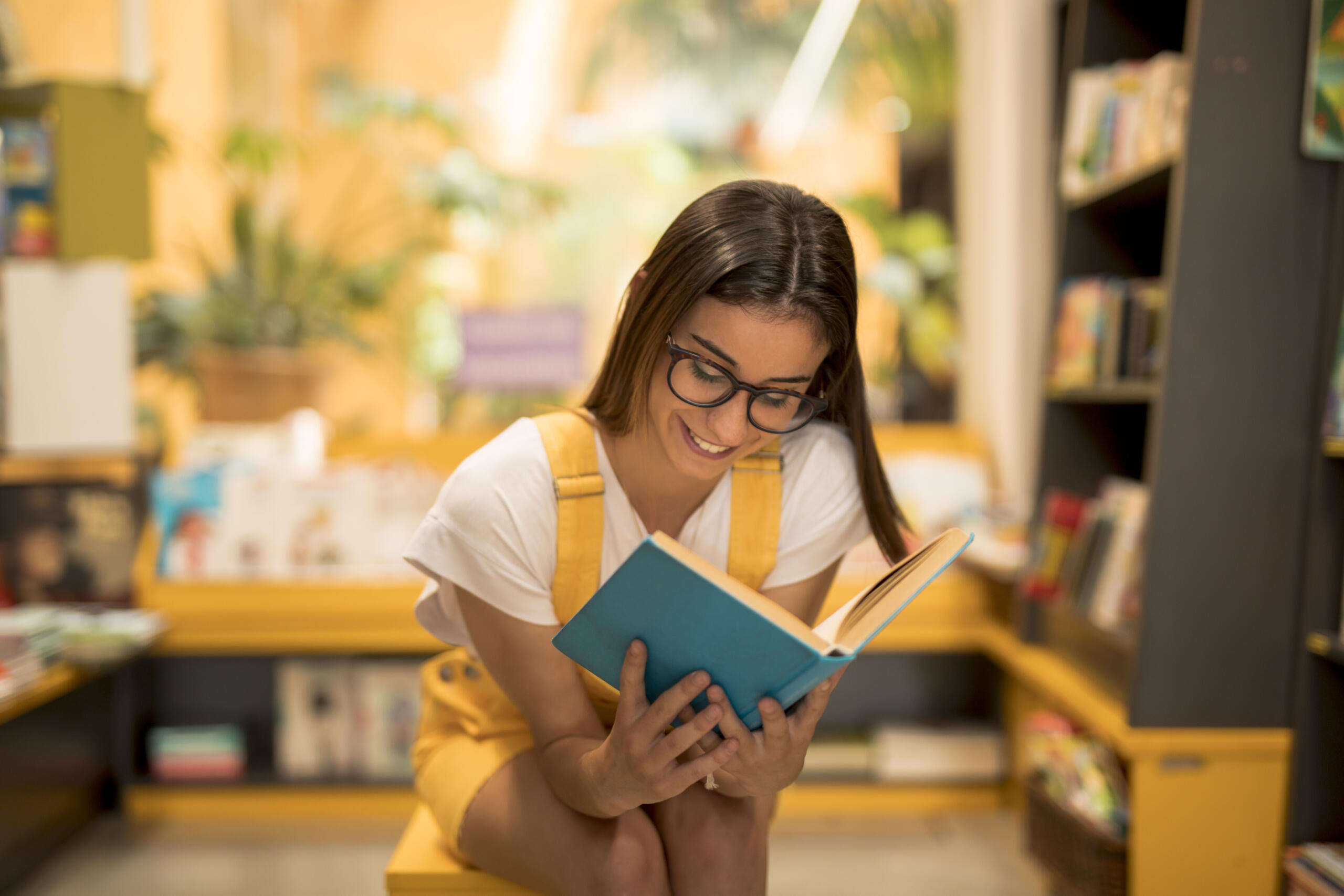 chica sonriente vestida de amarillo leyendo un libro celeste sentada en medio de una biblioteca en fondo desenfocado.