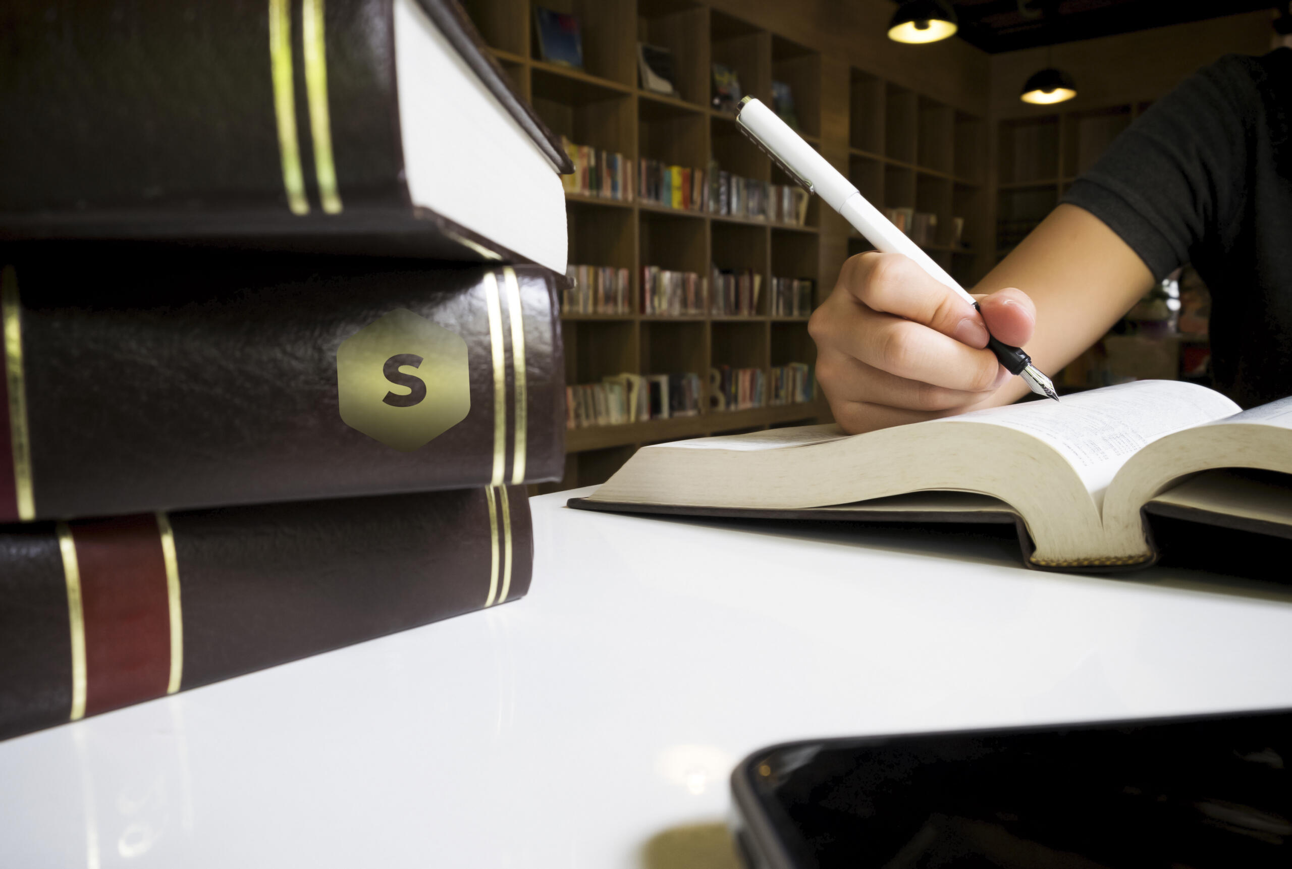 vista de una mano con libro abierto sujetando una pluma con libros apilados en la mesa con logo de Superprof en medio de la biblioteca