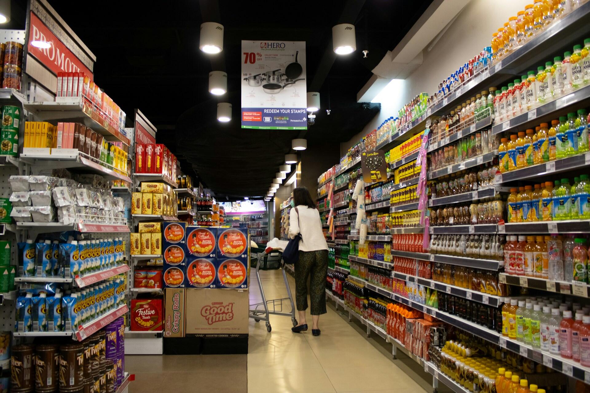 Frau schiebt Einkaufswagen durch Supermarkt mit vollen Regalen.