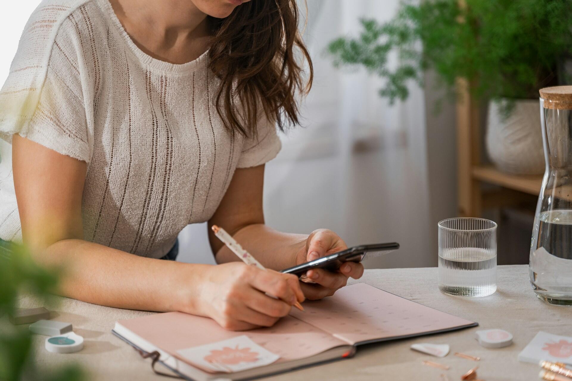 Frau sitzt an Schreibtisch mit Handy und Stift on der Hand vor Heft.